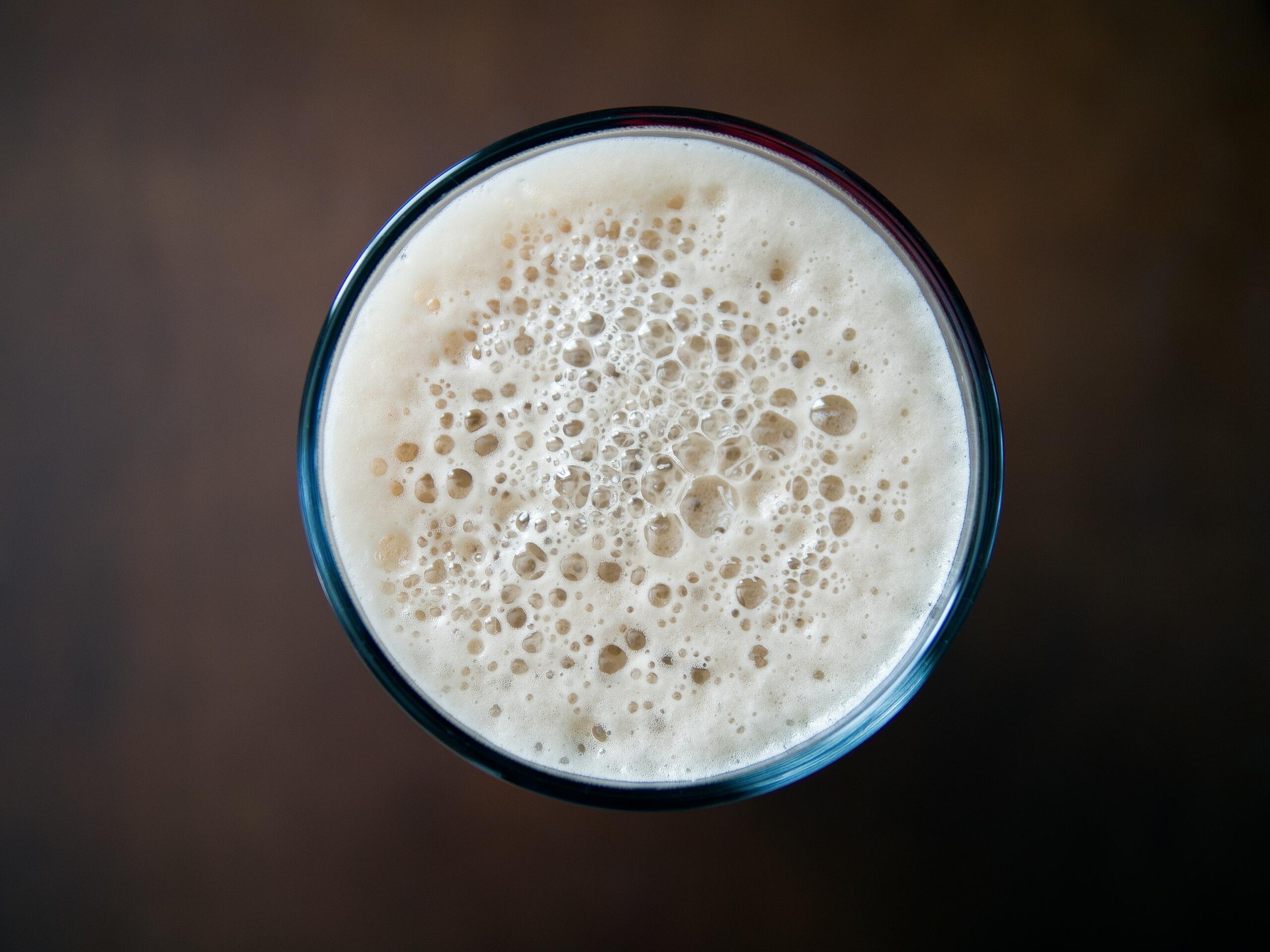 Top-down view of a glass filled with pale beer and a thick layer of white foam against a dark background.