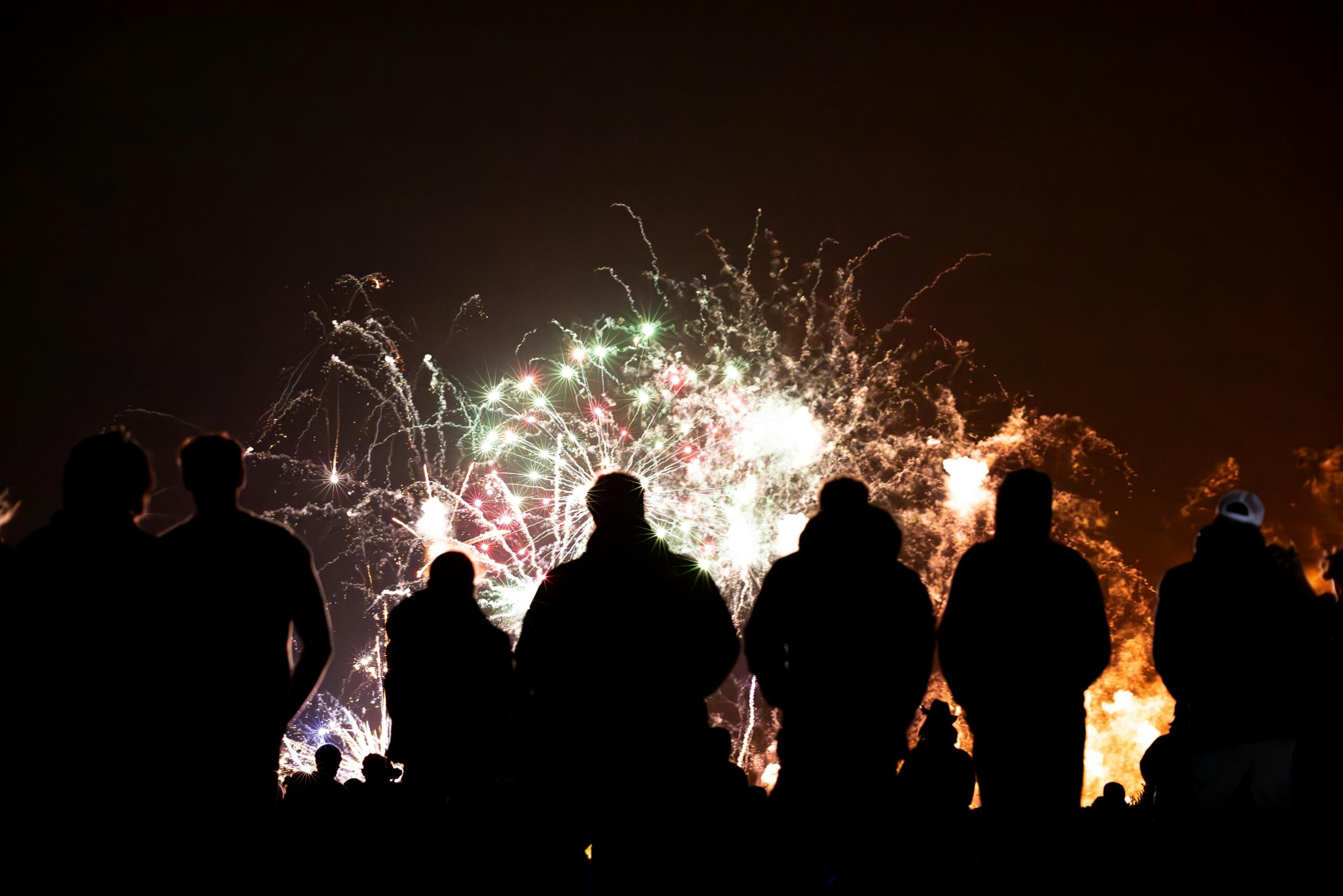 Silhouettes of spectators watching a bright fireworks display with white, red, green and orange bursts filling the night sky.