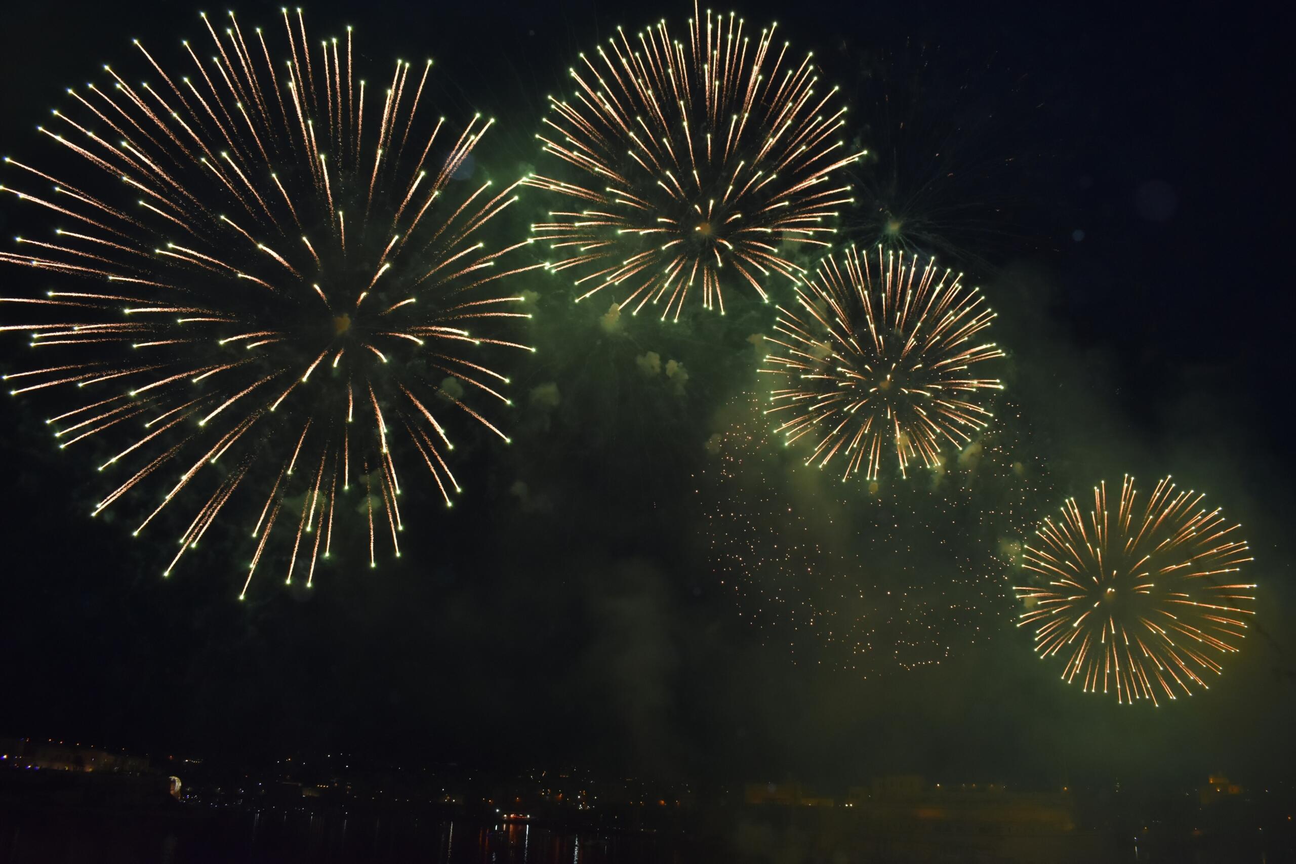 Multiple large golden fireworks bursting across the dark night sky above a waterfront in Malta.
