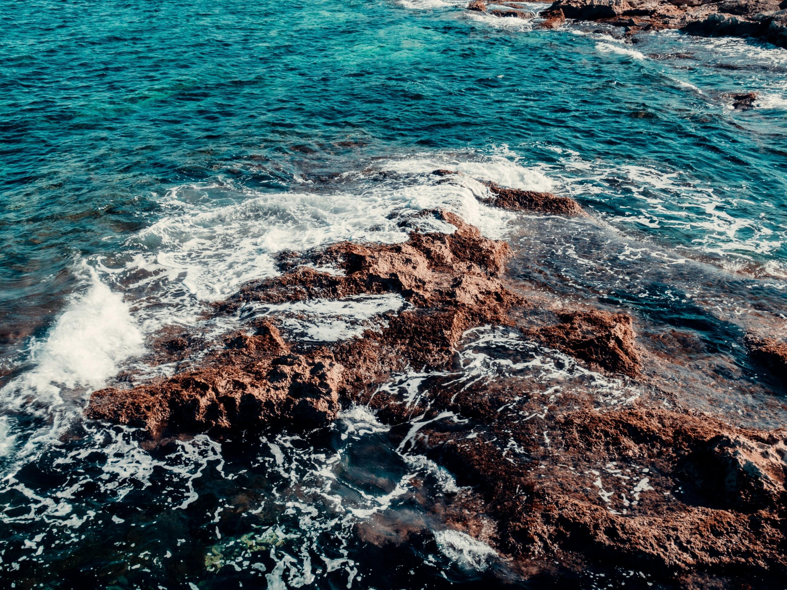 Turquoise seawater and white waves washing over jagged brown rocks along a rocky coastline.