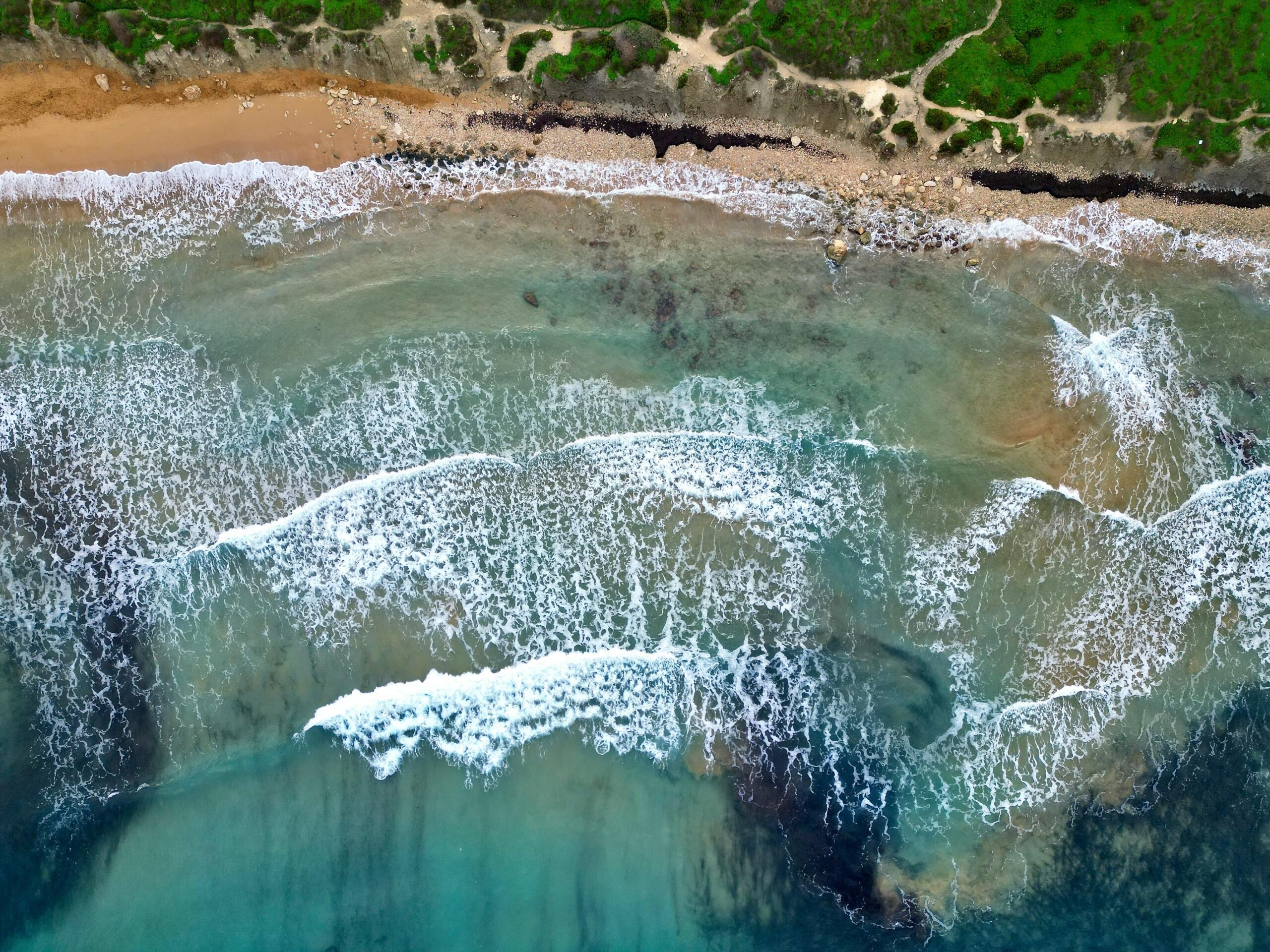 Aerial view of waves breaking along a sandy and rocky coastline bordered by green vegetation.