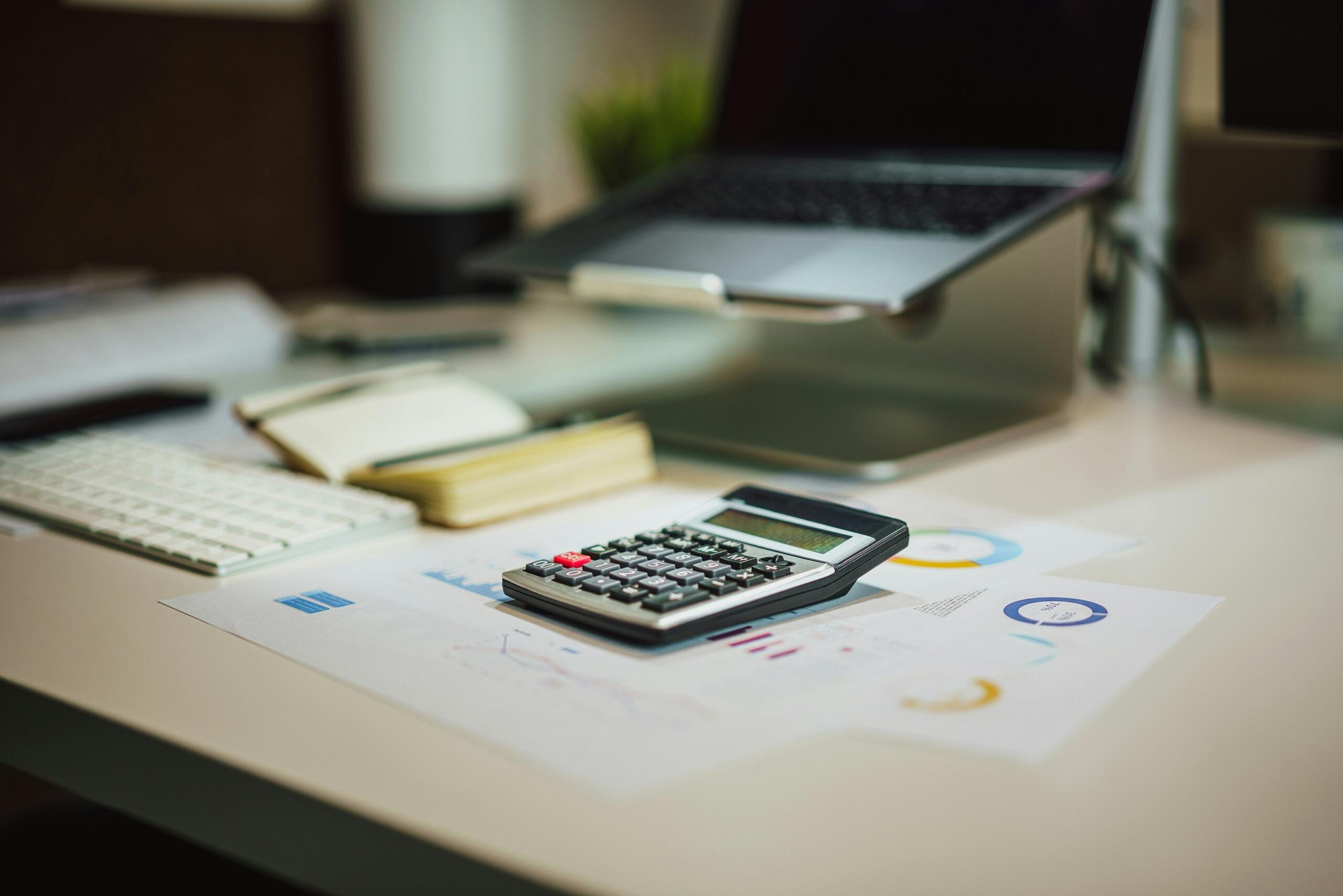 A calculator, notebook, pencil and pair of glasses lie on a wooden desk beside a laptop.