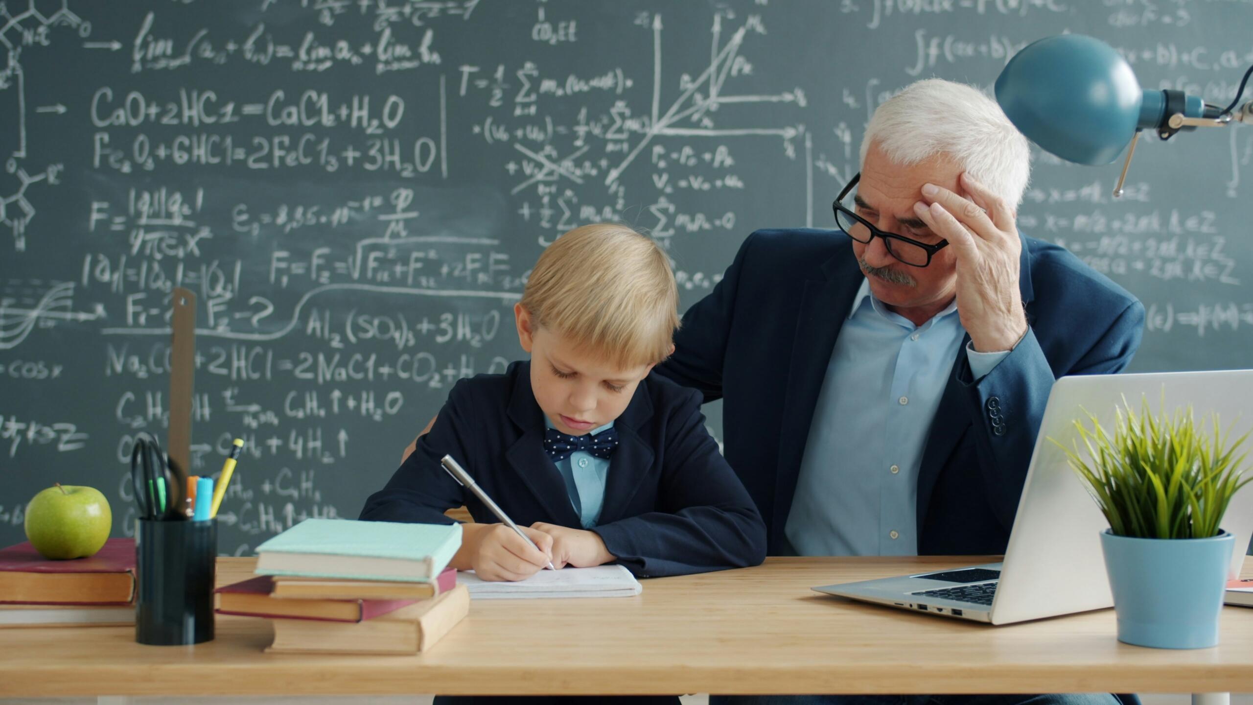 A tutor leans beside a young student who is writing at a desk, with a laptop and chalkboard in the background.