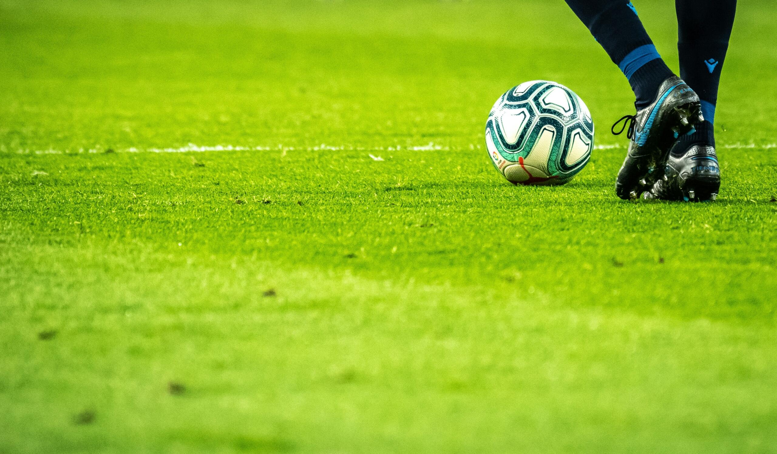 Close up of a football player's boot next to a football on a grass pitch.