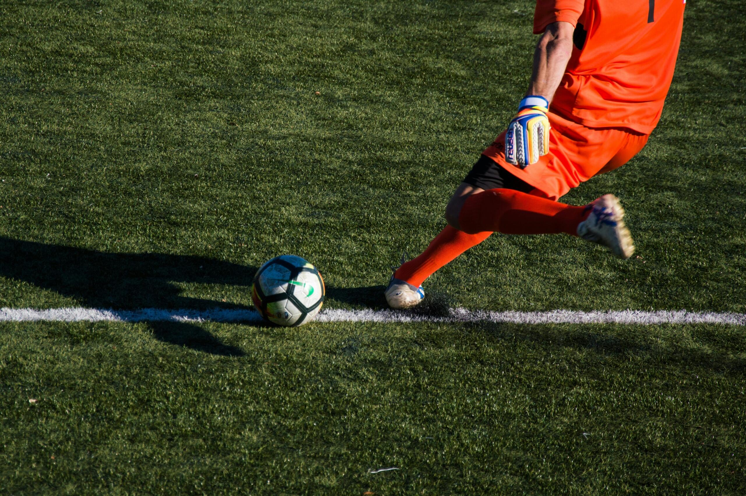 Goalkeeper in orange kit about to kick a football on a grass pitch.