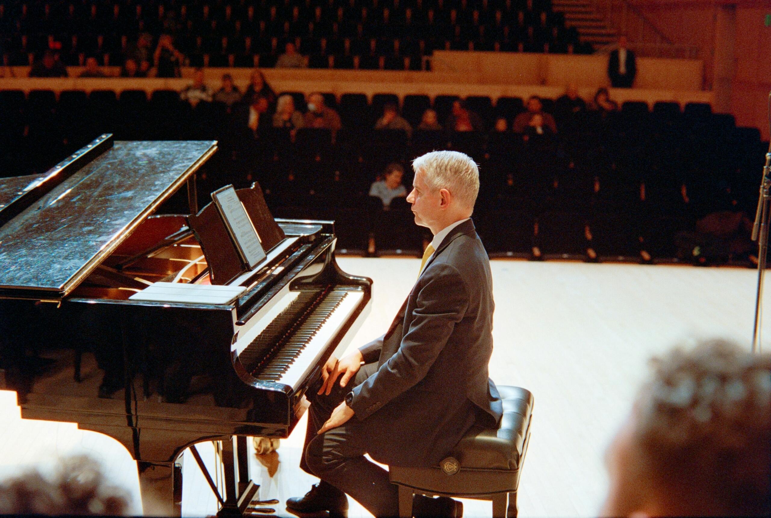 A pianist in a dark suit sits at a grand piano on stage with an audience in the background.