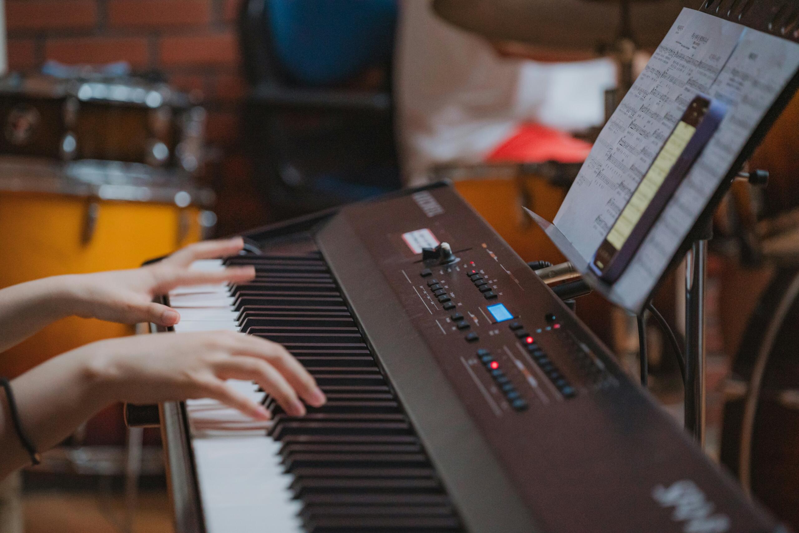 Two hands play a digital piano beside sheet music displayed on a music stand.