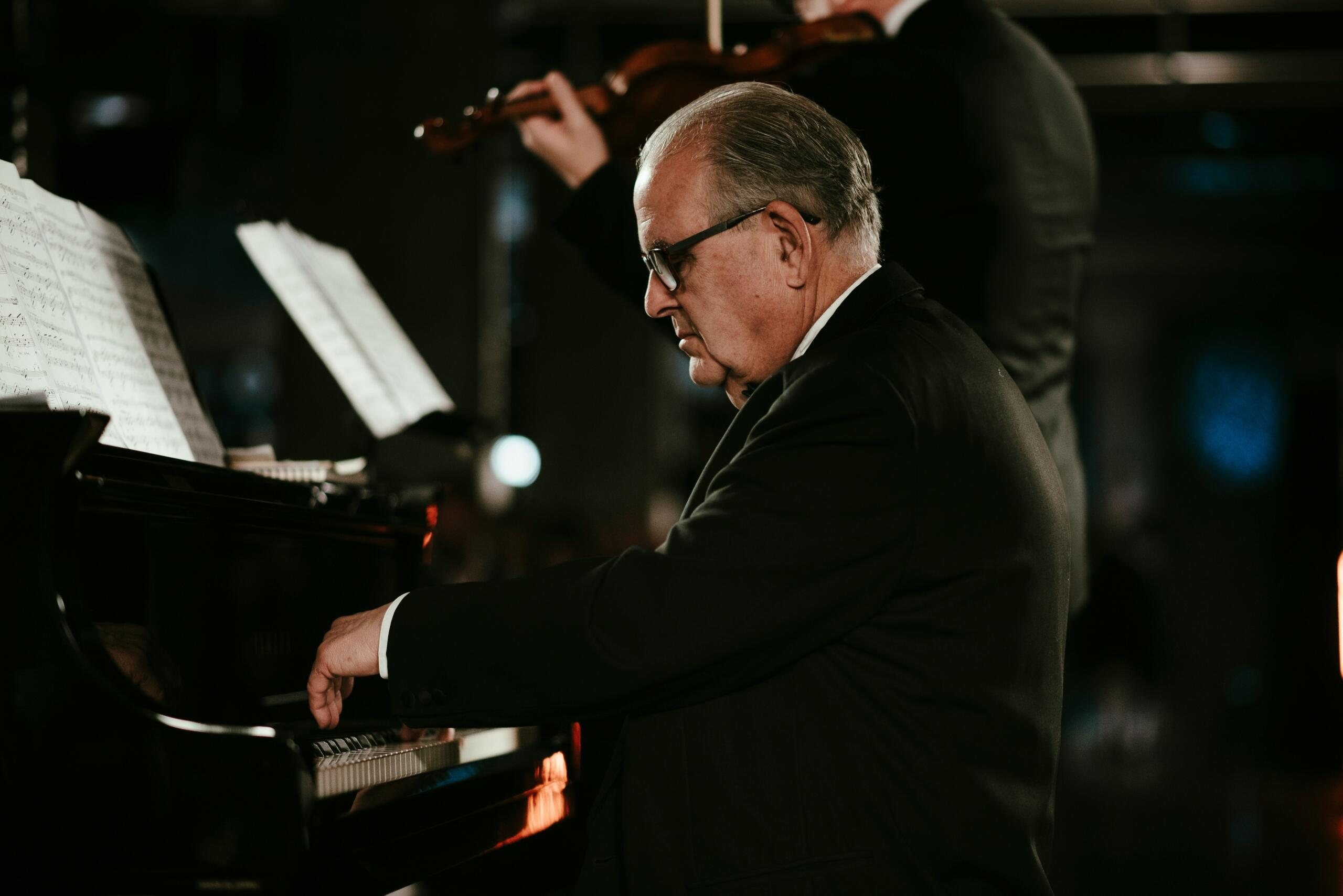 A pianist plays a grand piano while a violinist performs behind him.