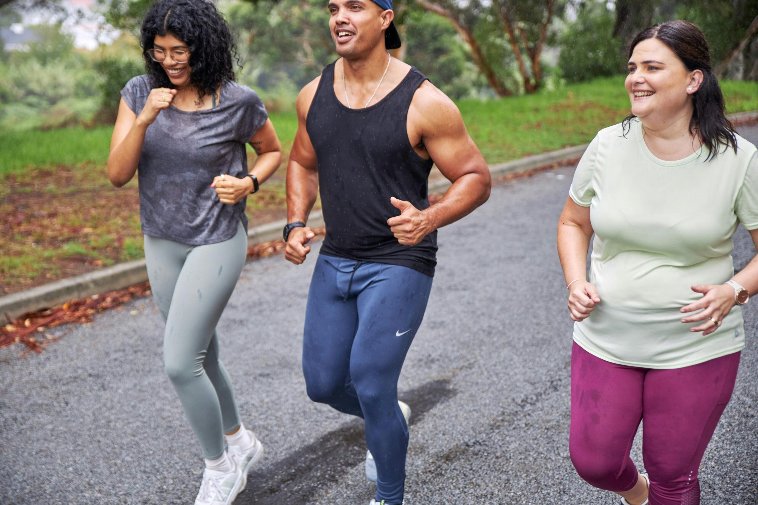 Three runners smiling as they train together on a wet road surrounded by trees and grass.
