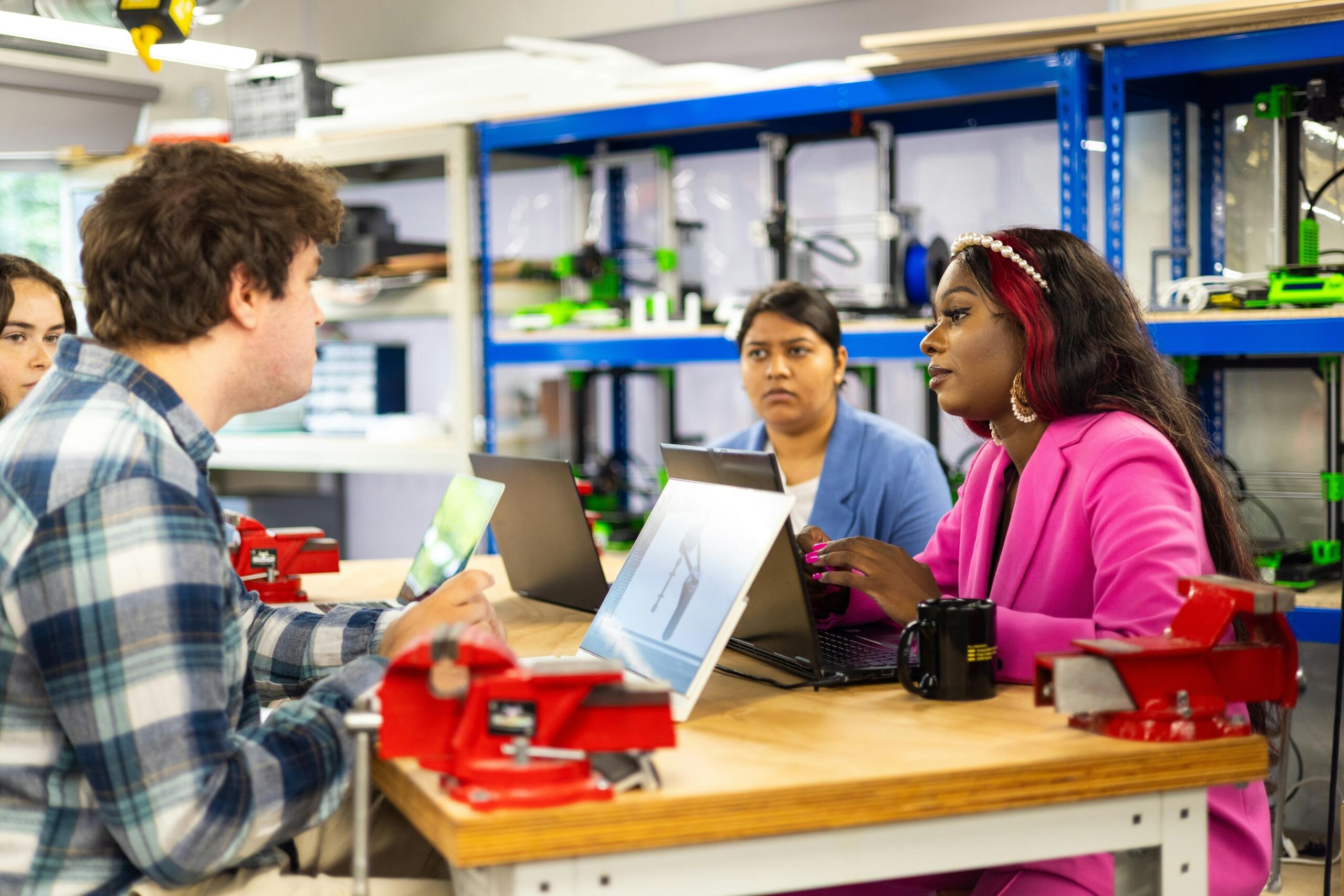 Diverse group of students working with laptops in a workshop environment with equipment on shelves