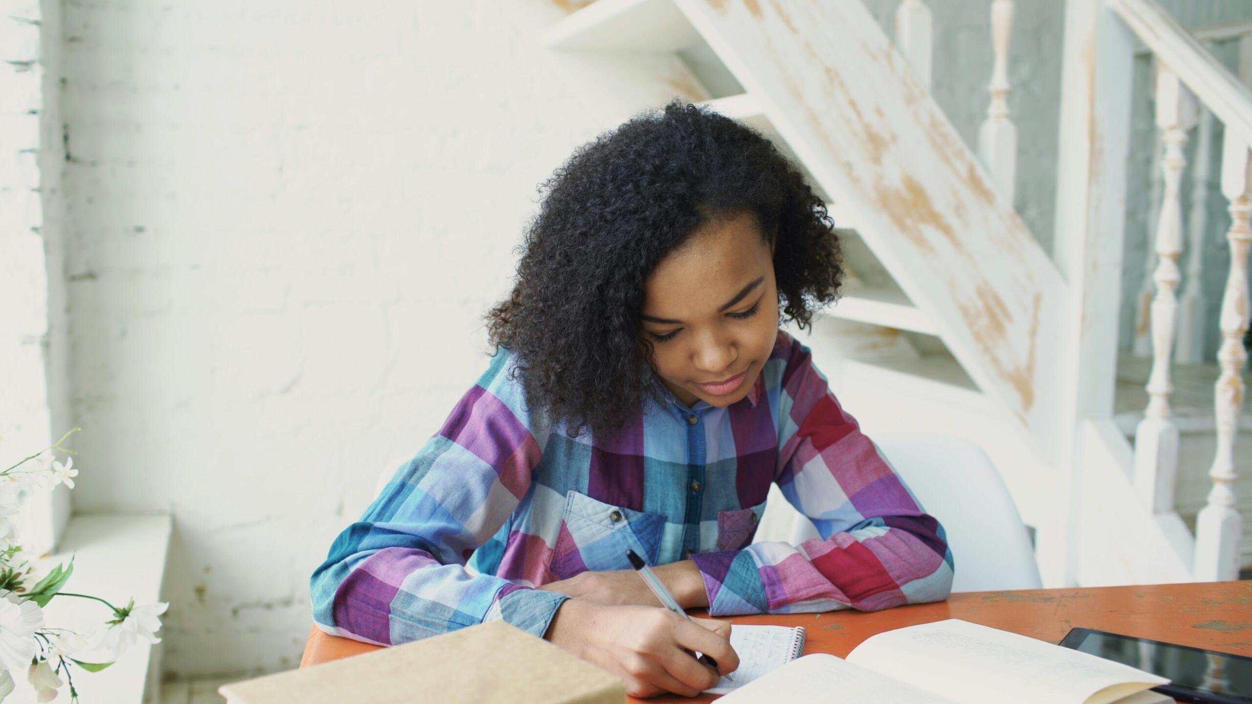 Young woman writing notes in a notebook at a desk with an open book and tablet