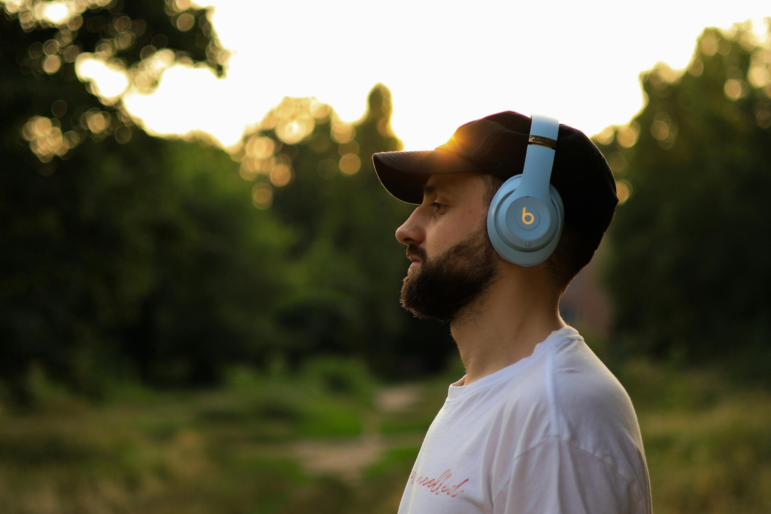 A man wearing blue wireless headphones and a black cap stands outside in a park at sunset, listening to music.