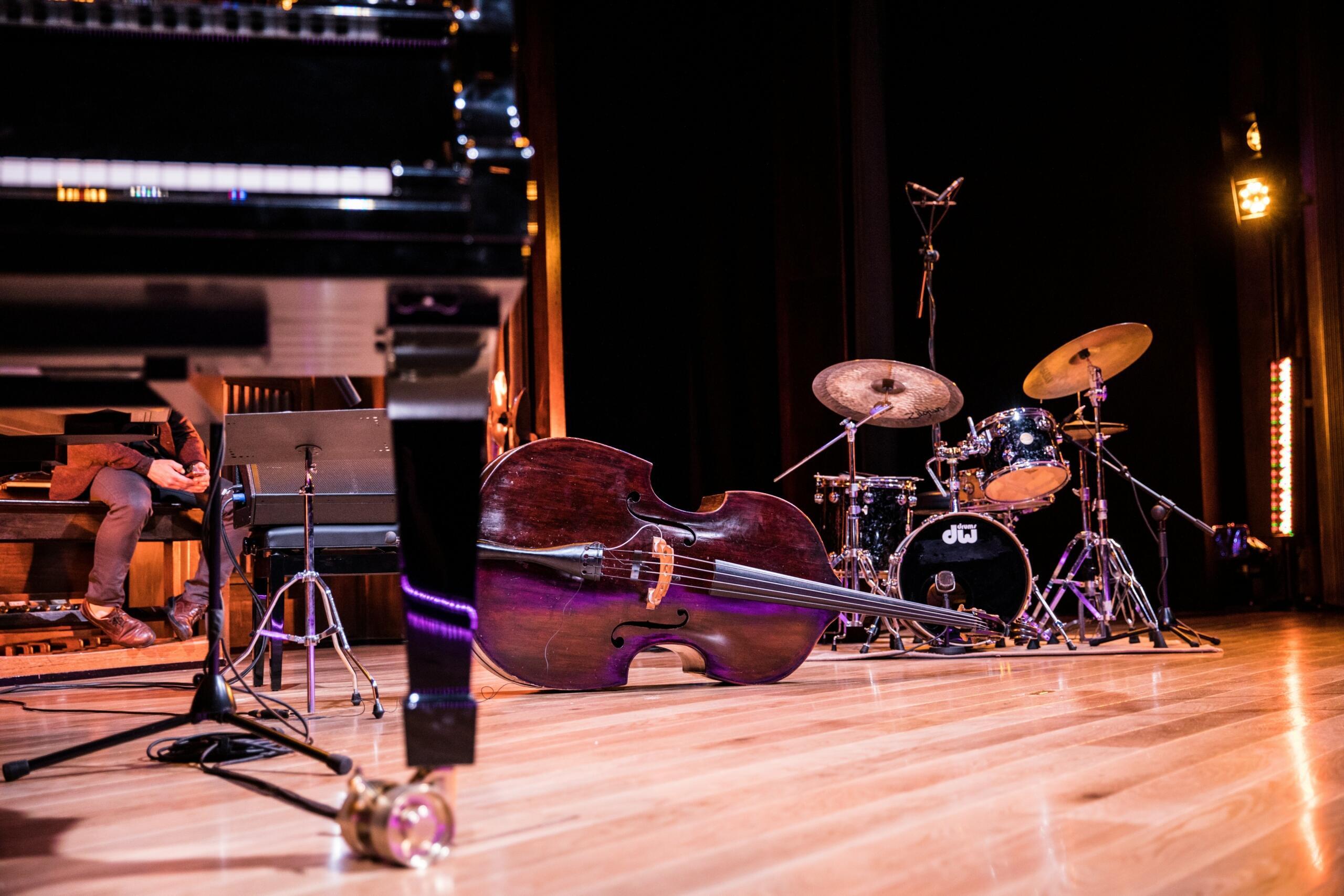 A stage setup featuring a double bass lying on the floor, a drum set, and part of a grand piano, ready for a jazz performance.