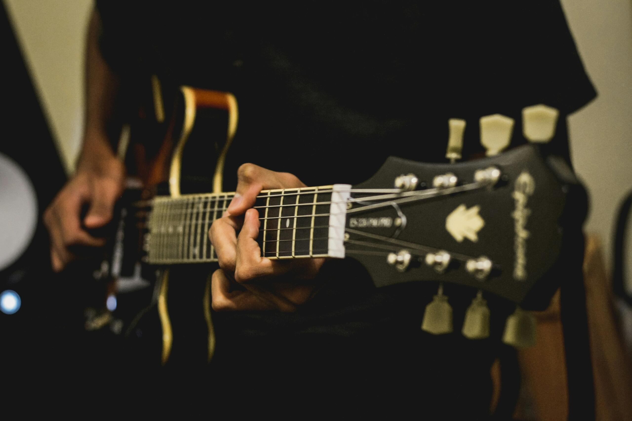 A person playing an electric guitar, focusing on their left hand pressing the strings on the fretboard and the headstock of the guitar.
