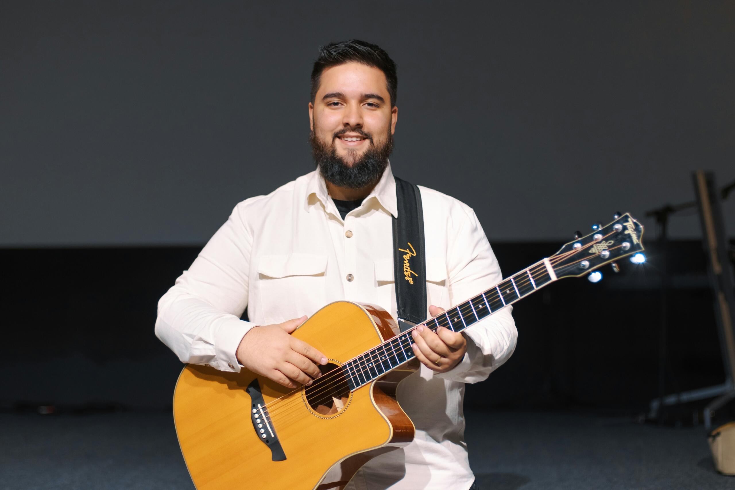 A smiling musician wearing a white shirt holds an acoustic guitar on stage, ready to perform.