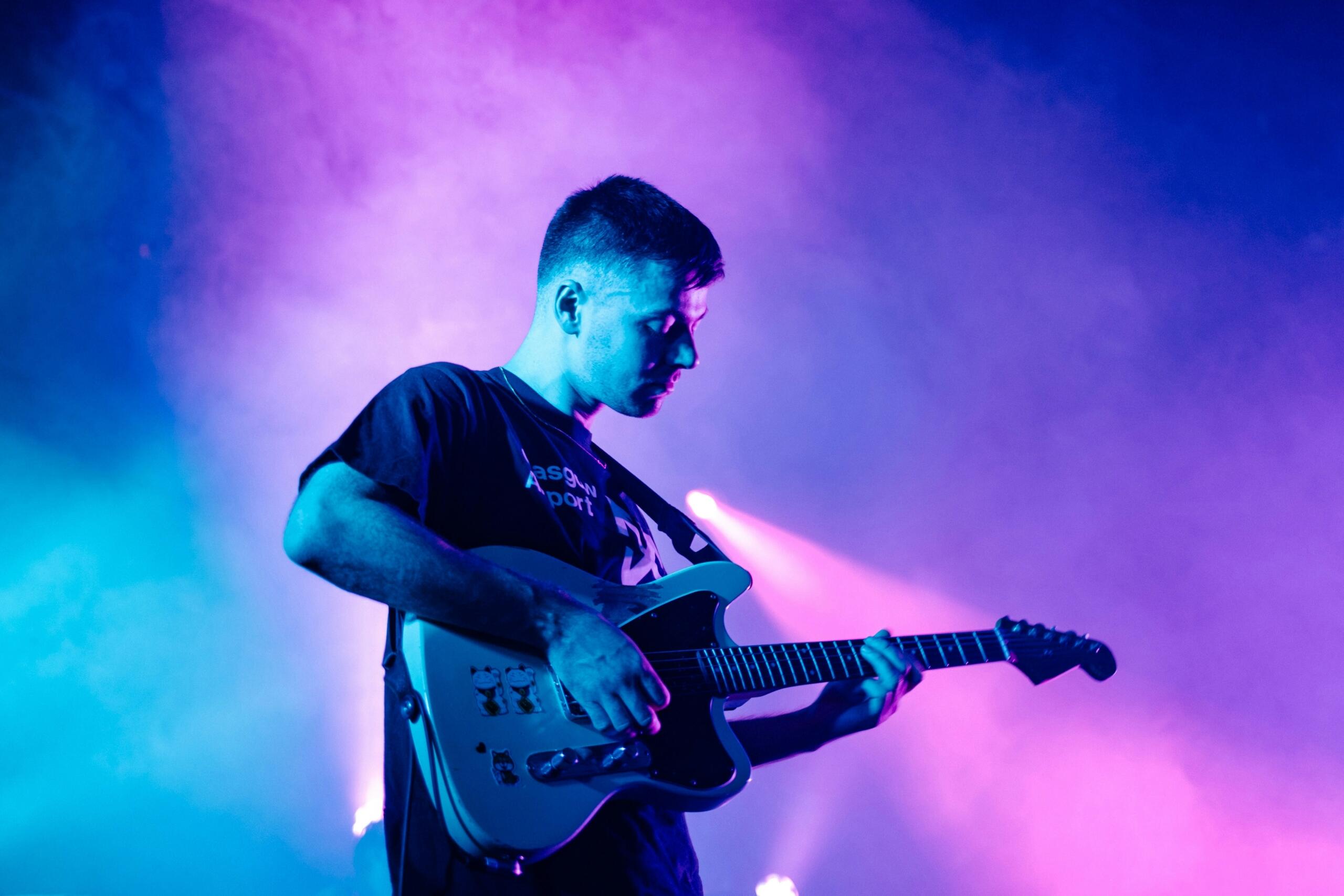 A guitarist performs on stage under vibrant blue and purple lights, focusing intently while playing an electric guitar.