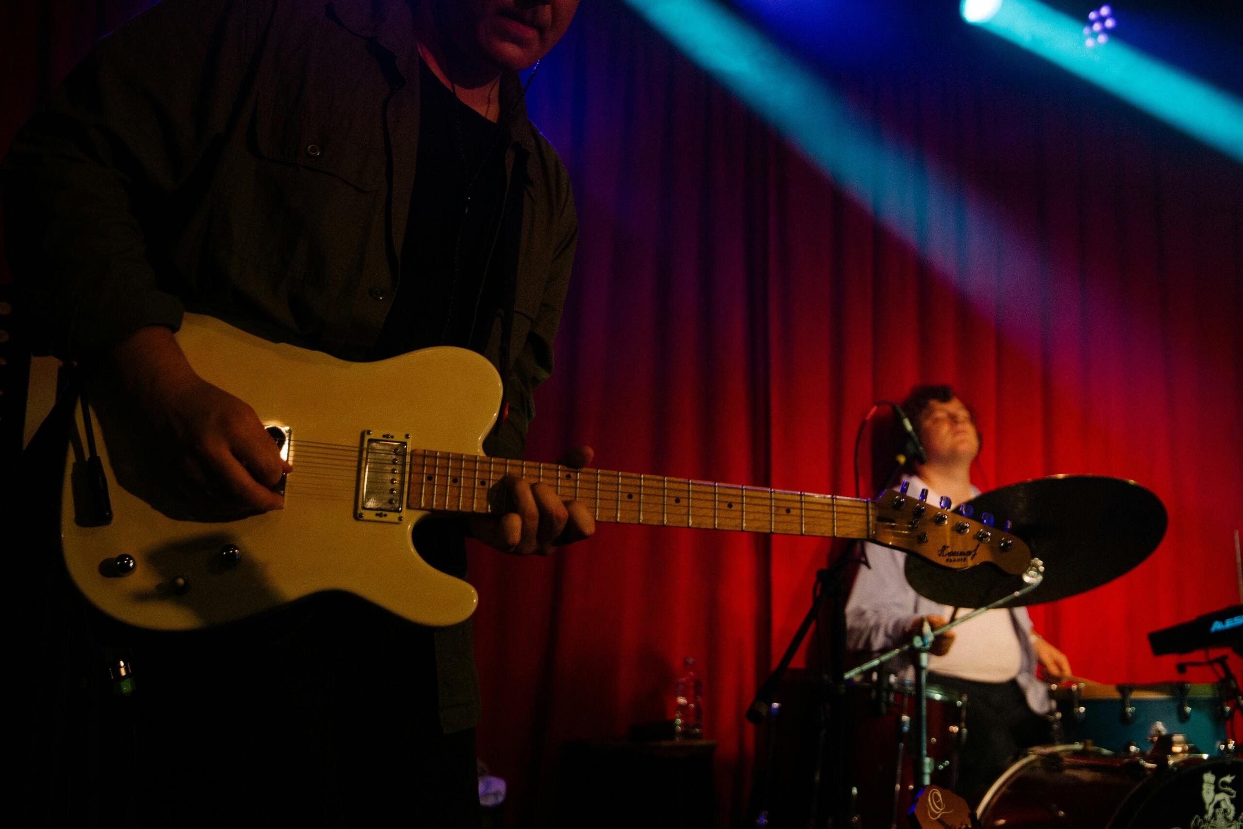 A musician playing an electric guitar on stage with a drummer performing in the background under stage lights and red curtains.