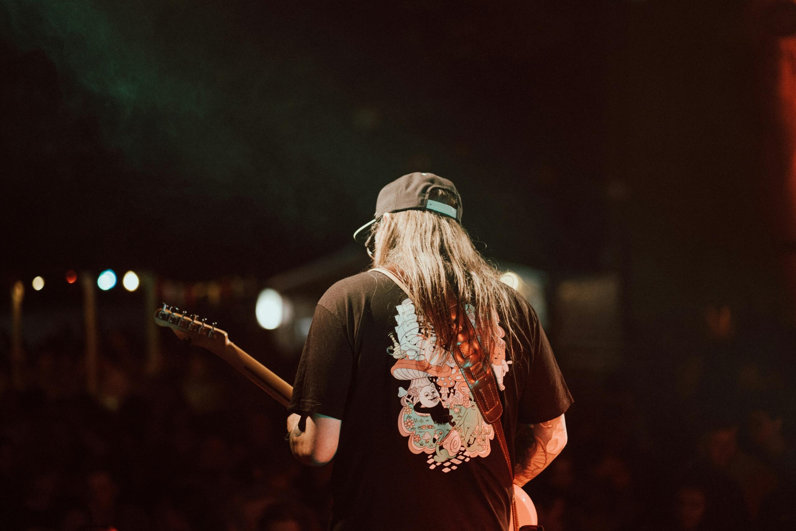 A musician with long hair and a black cap plays guitar on stage, viewed from behind, under warm lighting with a crowd in the background.