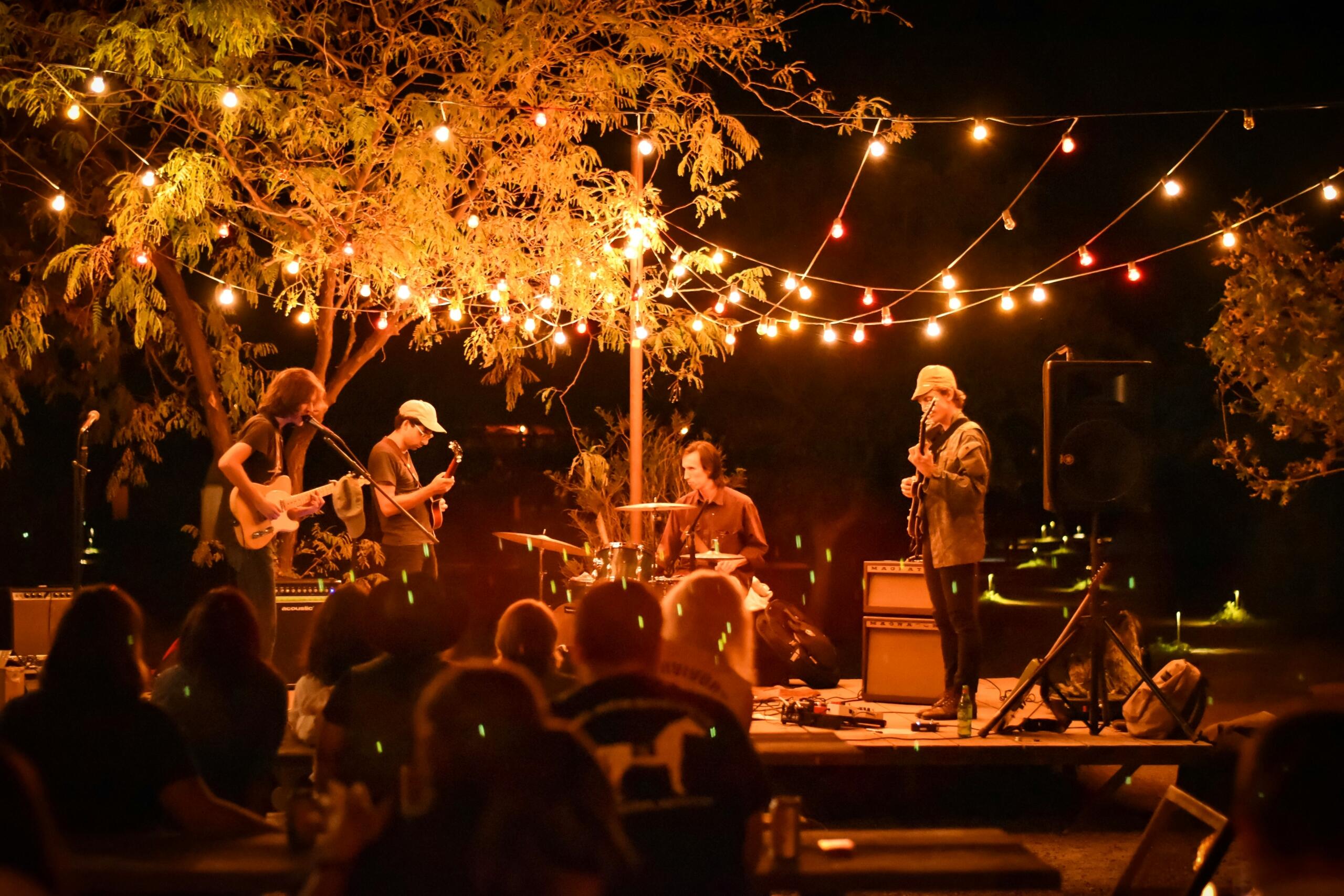 A live band performs outdoors at night under warm string lights, surrounded by trees and an audience seated at tables enjoying the music.
