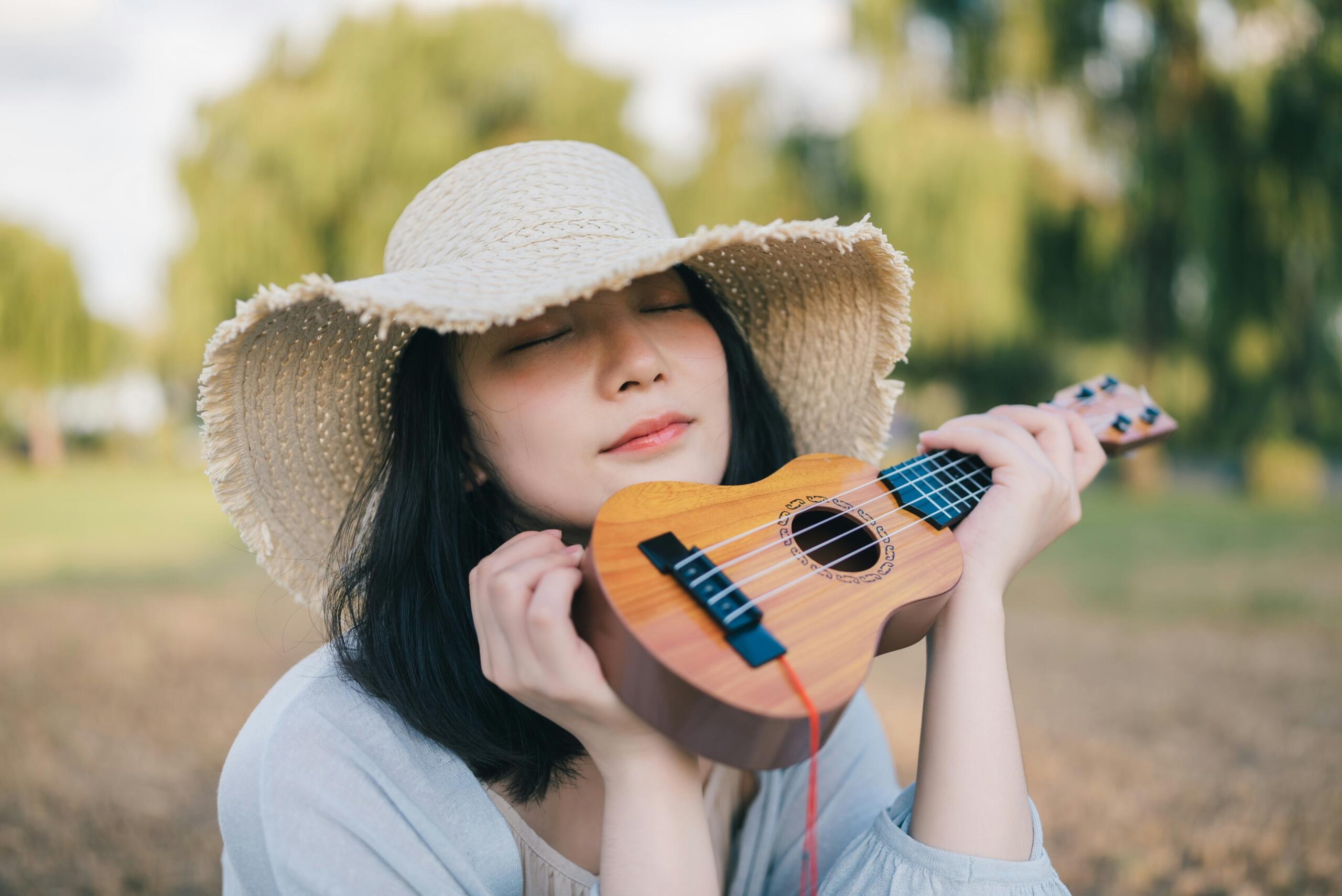 A woman wearing a wide-brimmed hat closes her eyes and smiles softly while holding a small ukulele close to her face in a peaceful outdoor setting.