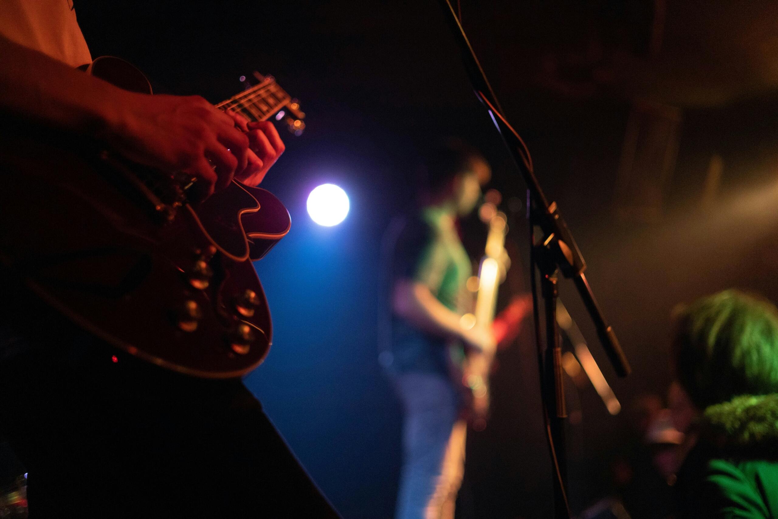 A close-up of a guitarist playing an electric guitar on stage, with another musician blurred in the background under concert lighting.