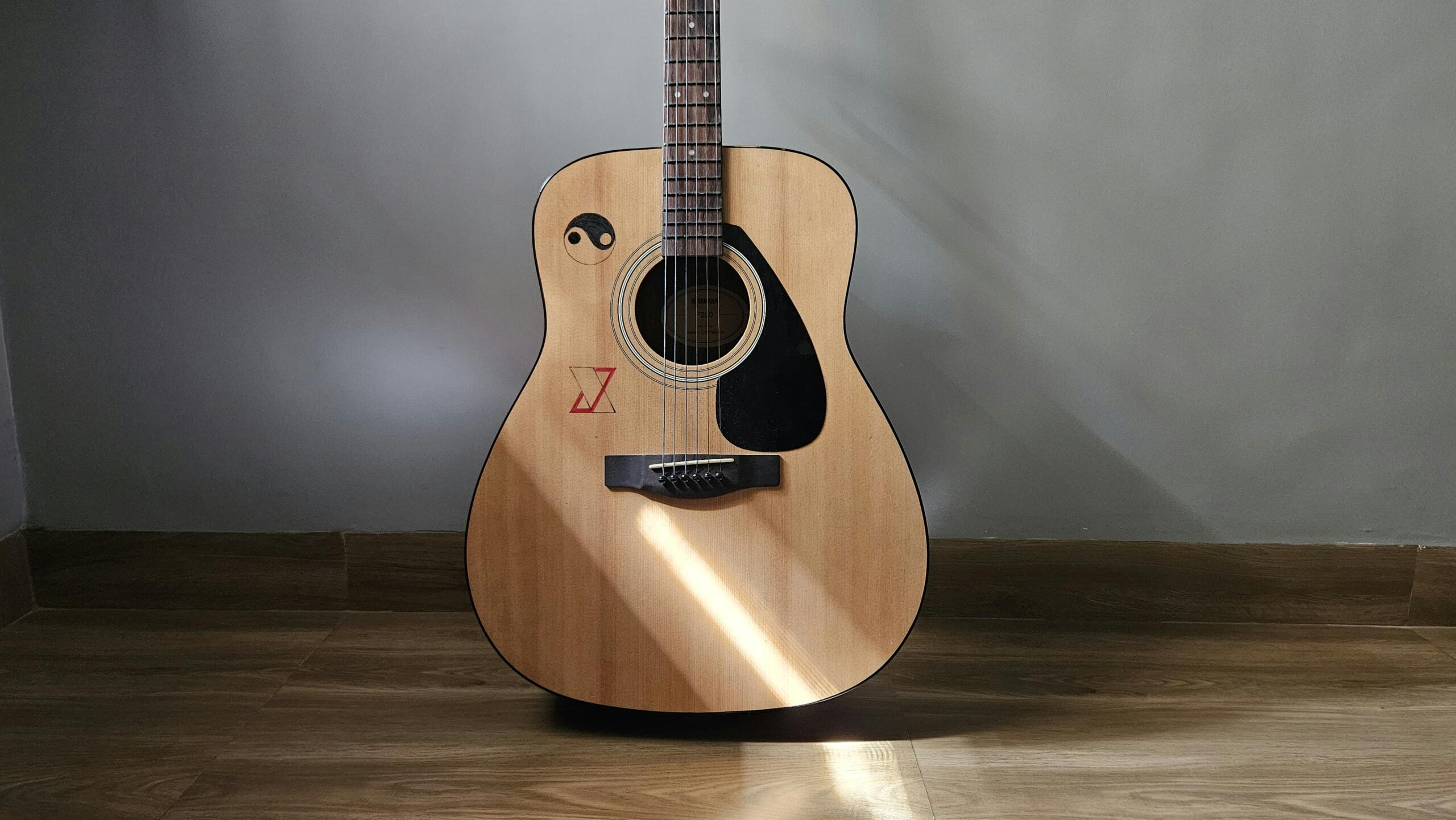 An acoustic guitar stands upright on a wooden floor with sunlight shining across its body.