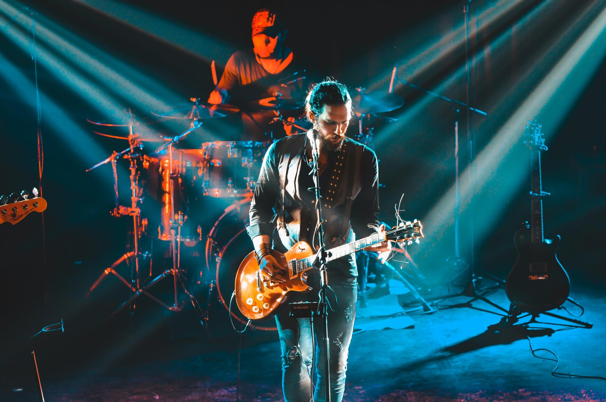 A guitarist and drummer perform on stage under dramatic lighting with beams of blue and orange light.