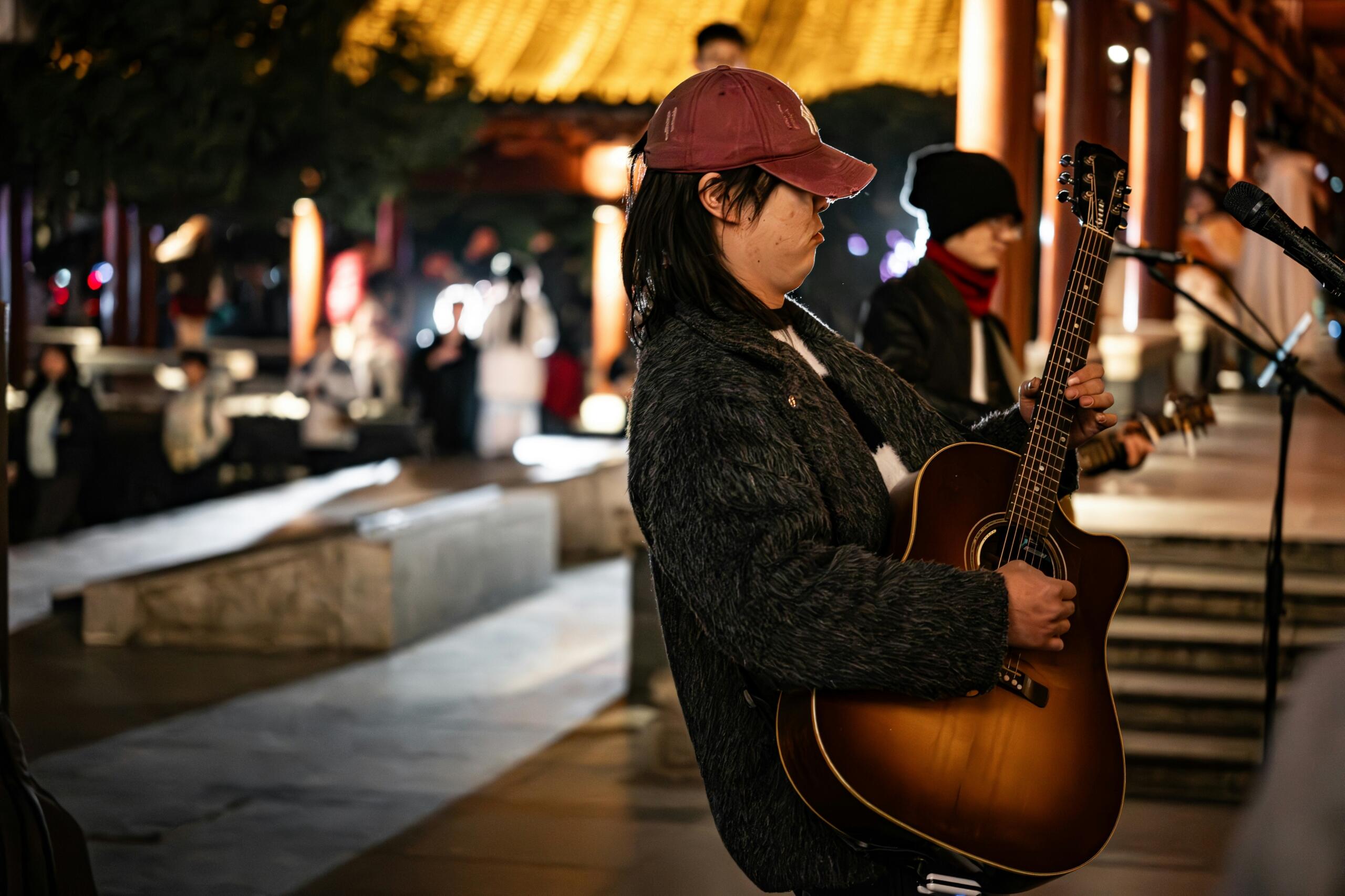 A street musician wearing a red cap and dark jacket plays an acoustic guitar during a nighttime performance, with a softly lit crowd in the background.