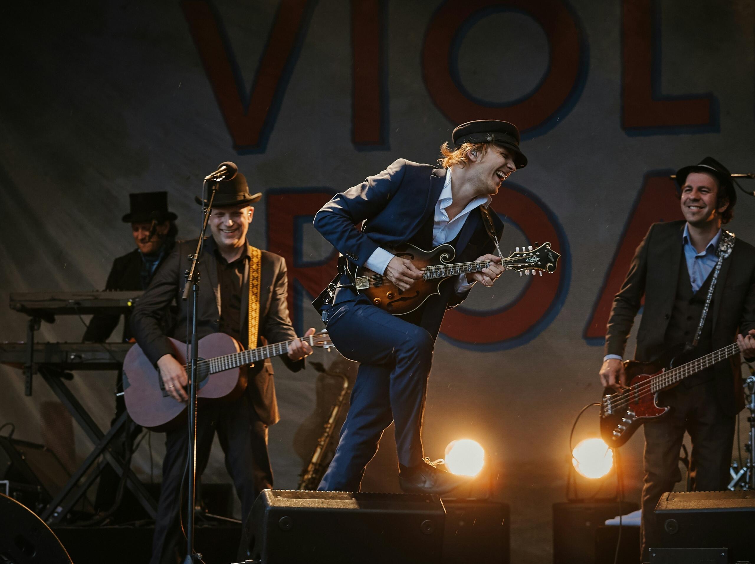 A lively band performs on stage with guitars and bright spotlights, with the lead musician energetically playing a mandolin while others smile and play along.