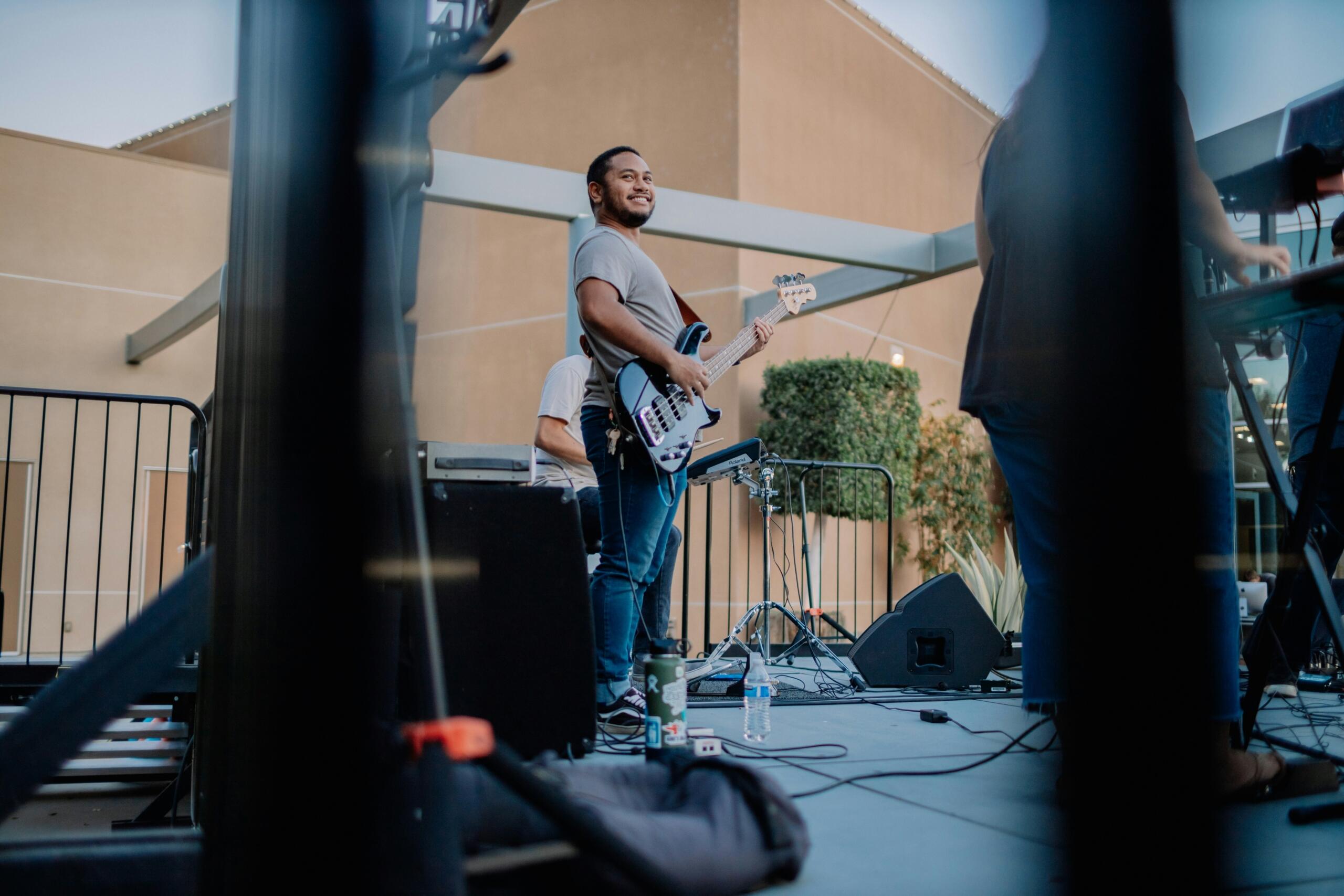 A smiling musician plays an electric bass guitar during an outdoor performance with other band members.