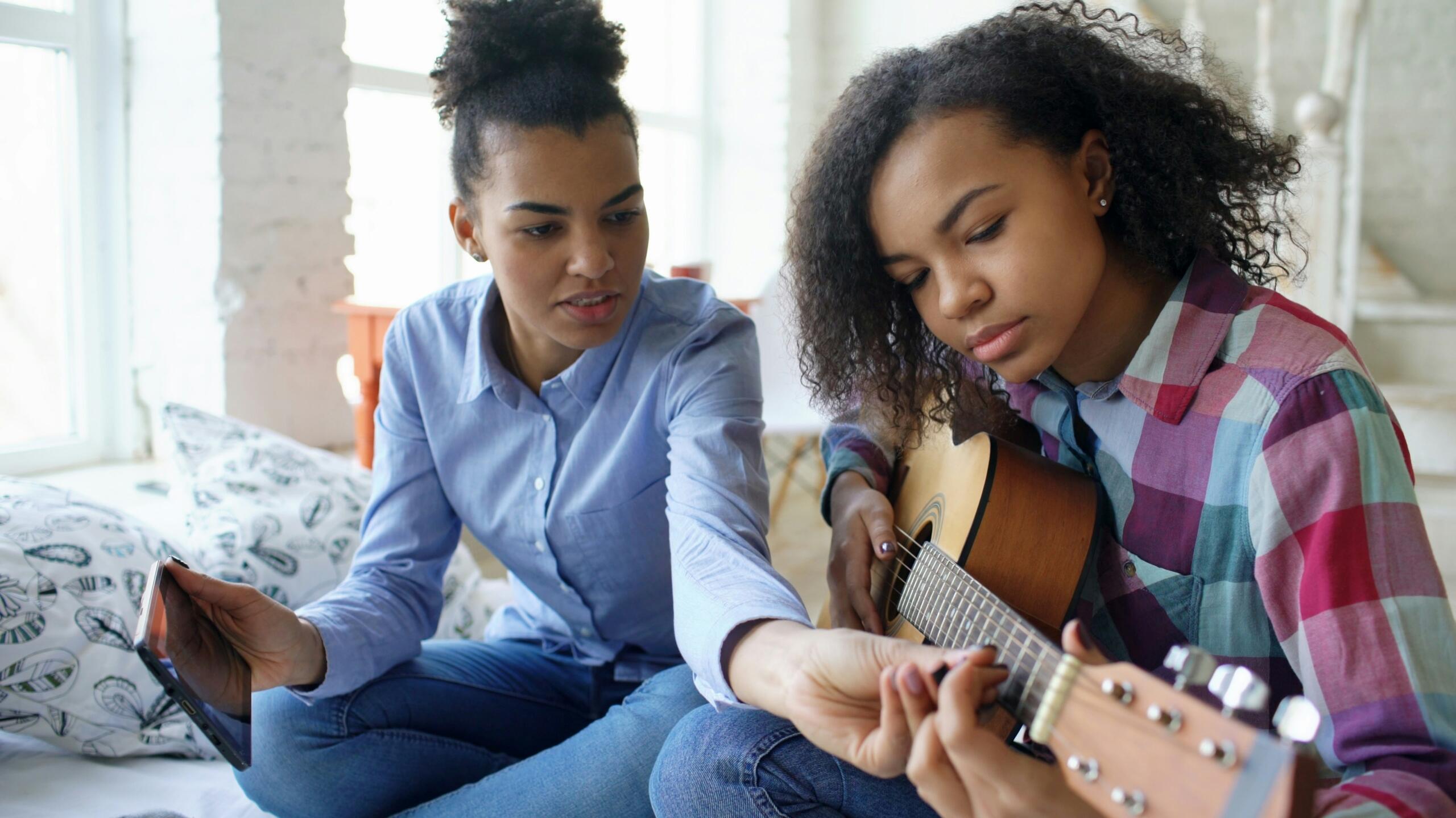 A woman teaches another woman how to play guitar, adjusting her fingers on the fretboard while holding a tablet in a bright room.