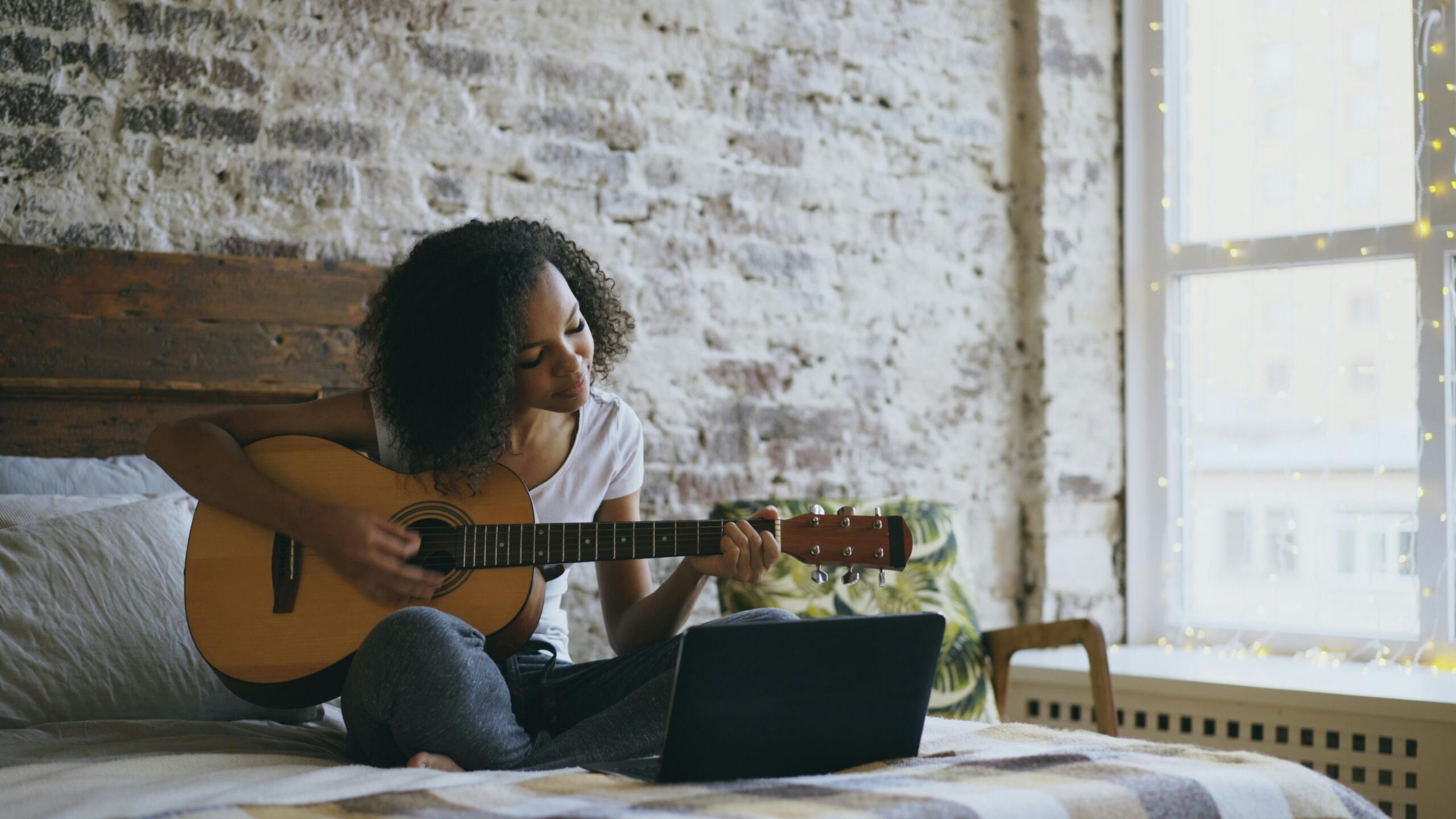 A woman sitting on a bed playing an acoustic guitar while looking at a laptop near a window with string lights.