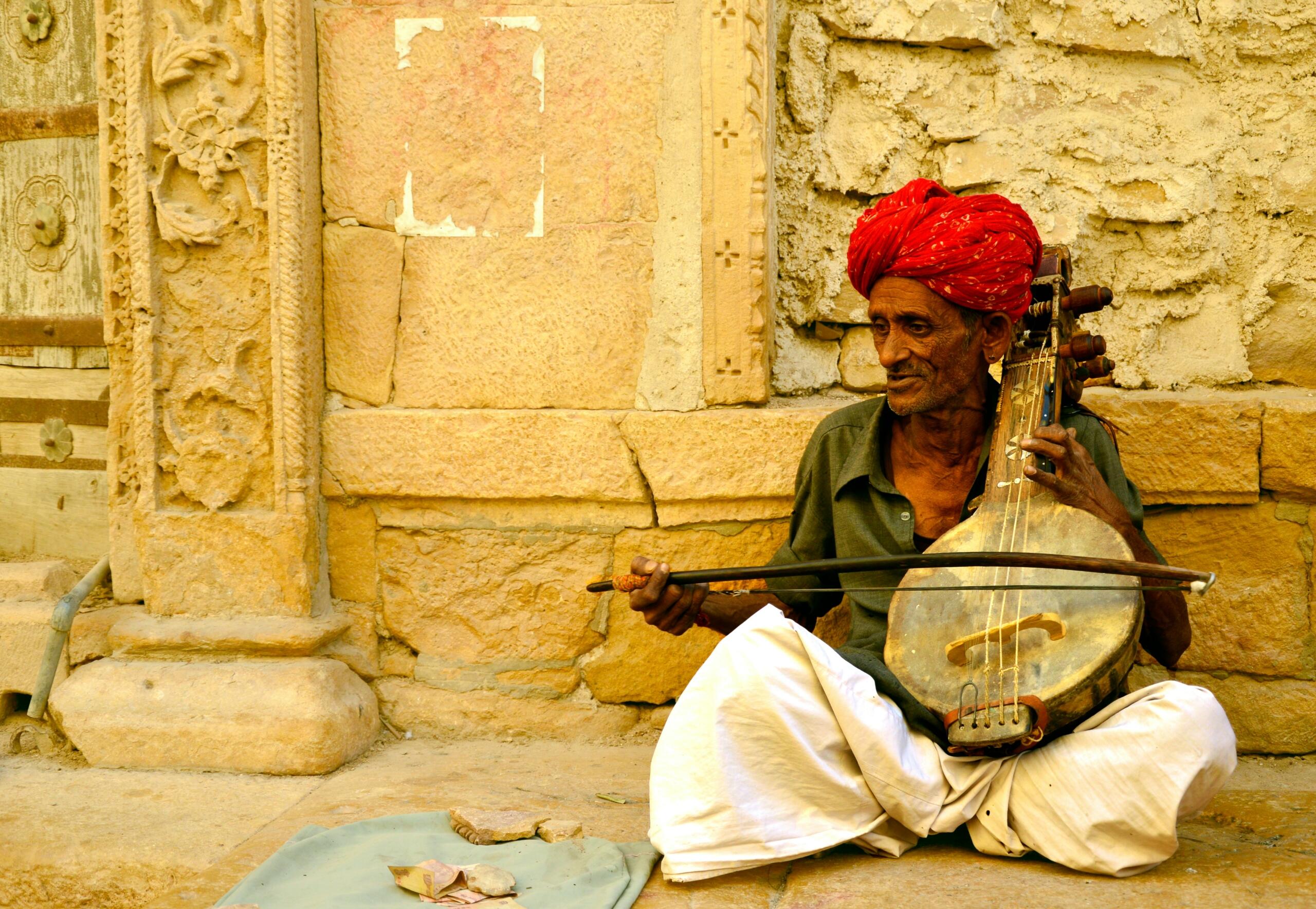 An elderly man wearing a red turban and white dhoti plays a traditional Indian string instrument with a bow while sitting against a sandstone wall.
