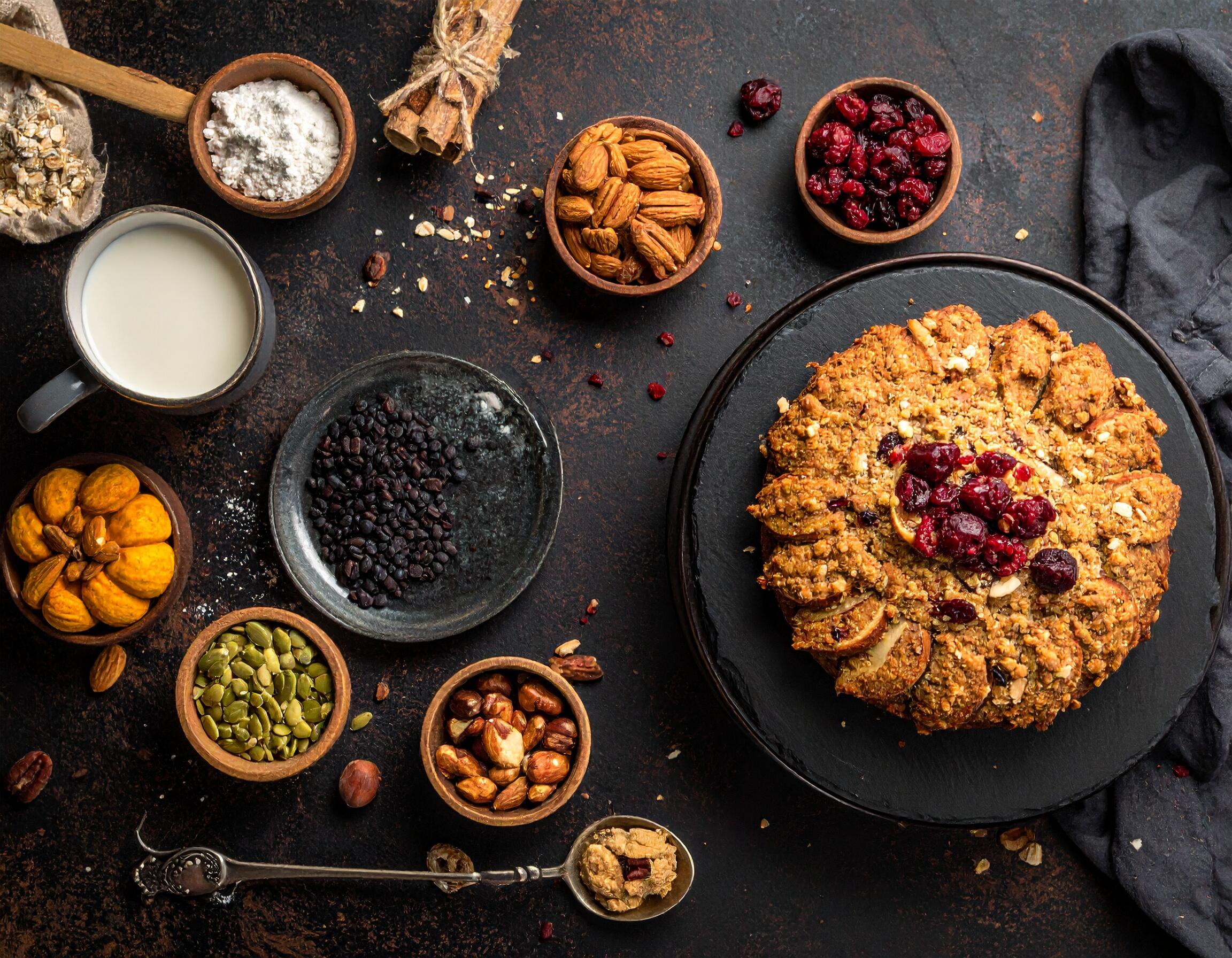 A beautifully arranged vegan baked dessert topped with cranberries, surrounded by baking ingredients like flour, nuts, seeds, oats, and a cup of plant-based milk on a dark rustic surface.