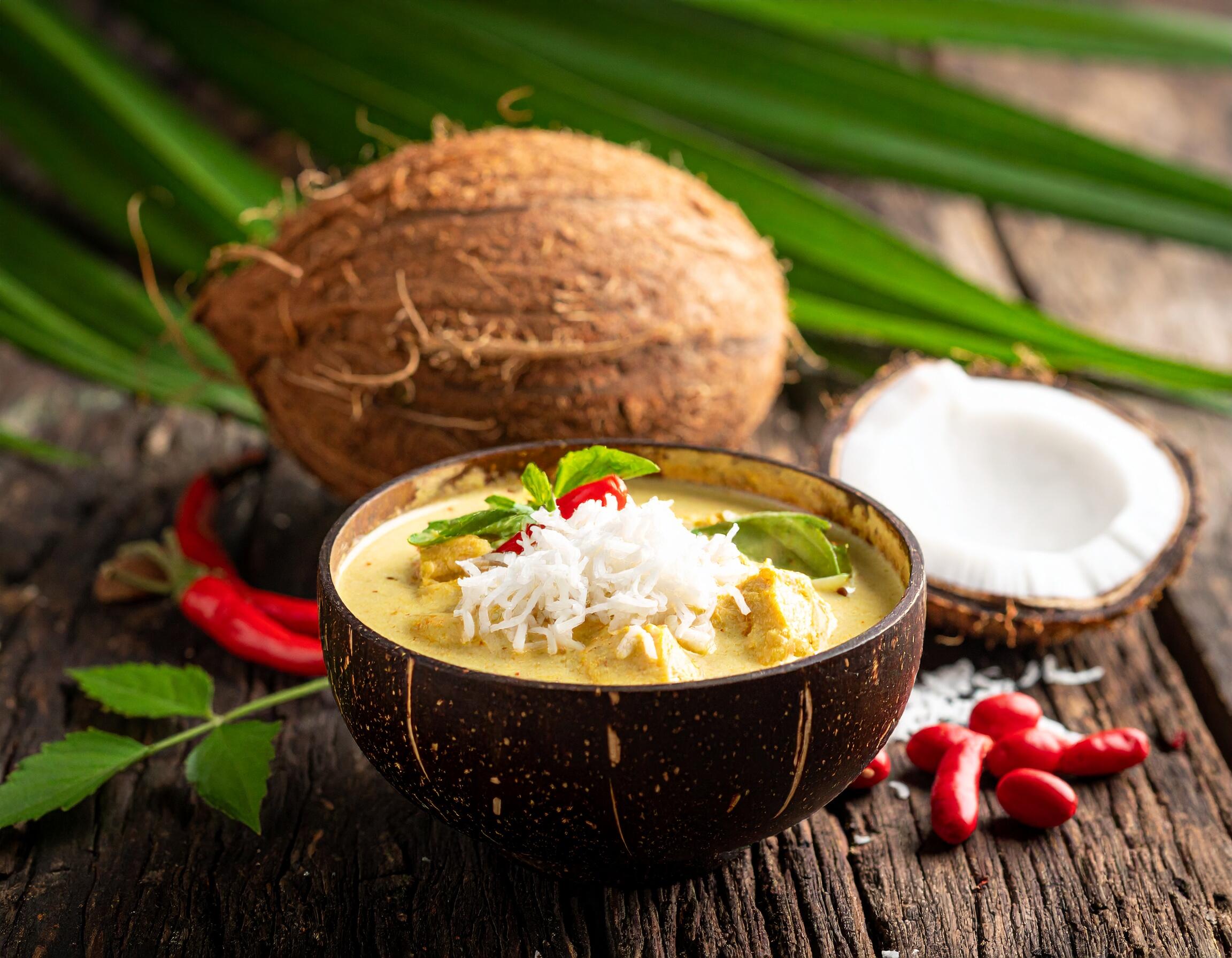 A bowl of vegan coconut curry garnished with shredded coconut, red chili, and fresh herbs, served in a coconut shell with coconuts and chili peppers in the background.