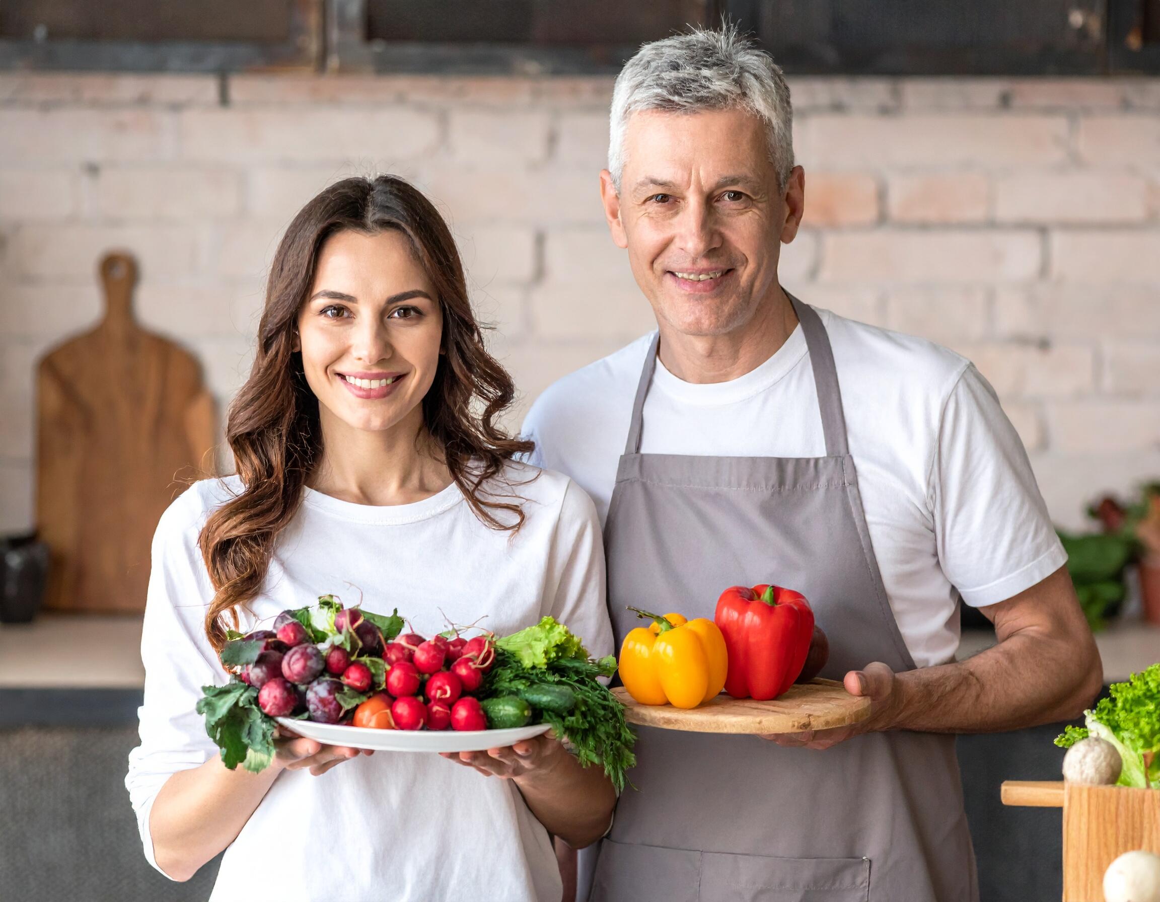 A smiling man and woman holding plates of fresh vegetables, including radishes, lettuce, and bell peppers, promoting vegan living in a bright kitchen.