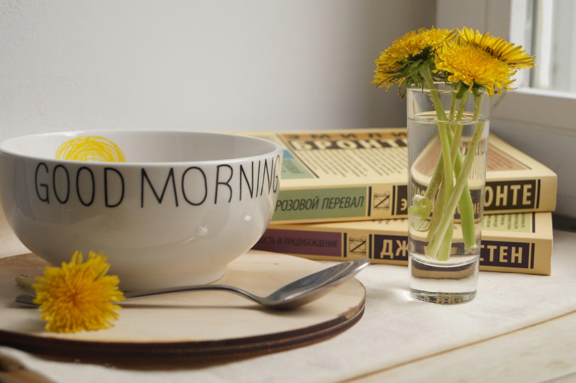 A white bowl with Good Morning text beside dandelions in a glass and a stack of books
