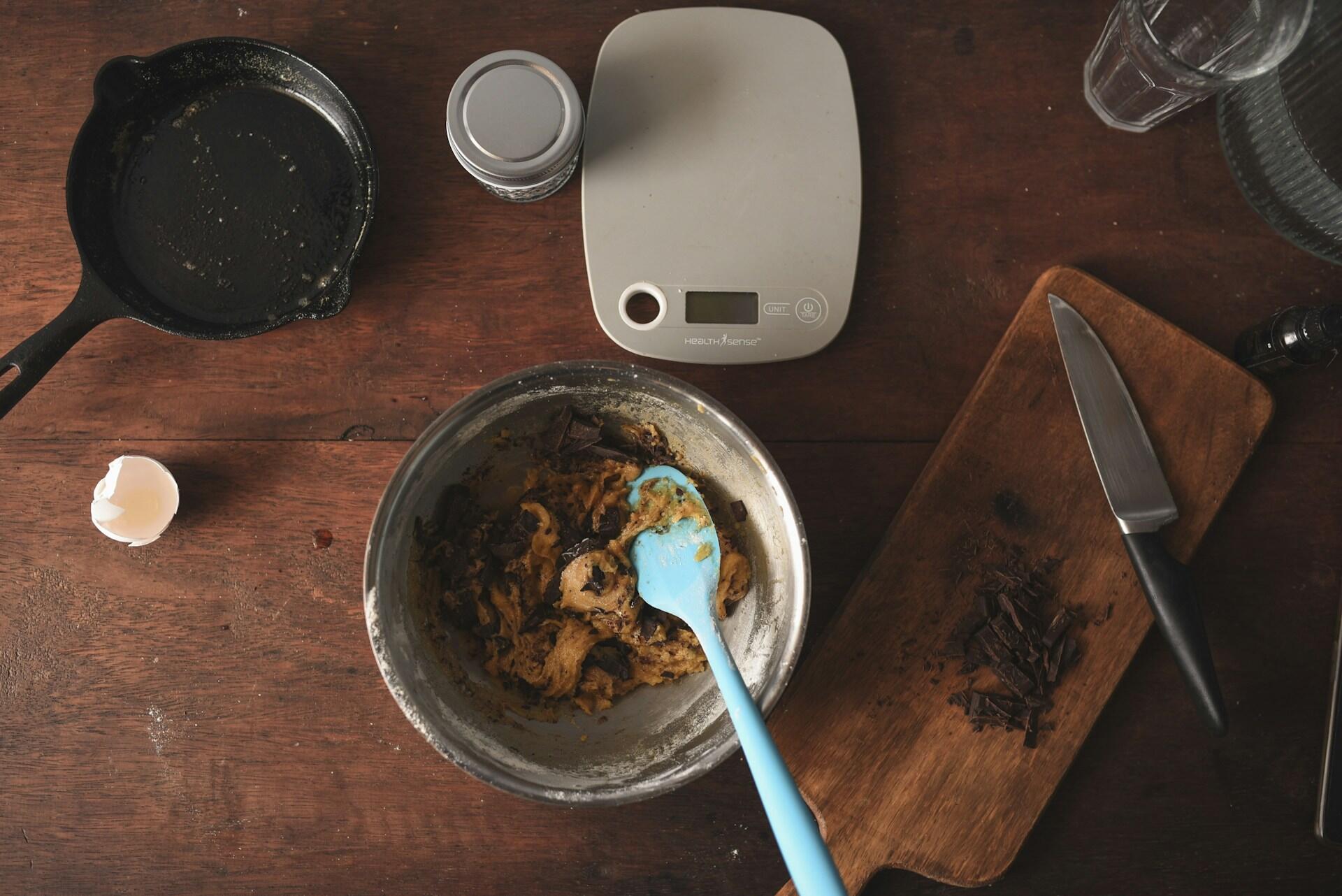 A cozy kitchen scene with cookie dough in a metal bowl, a blue spatula, chopped chocolate on a wooden board, and baking tools nearby.