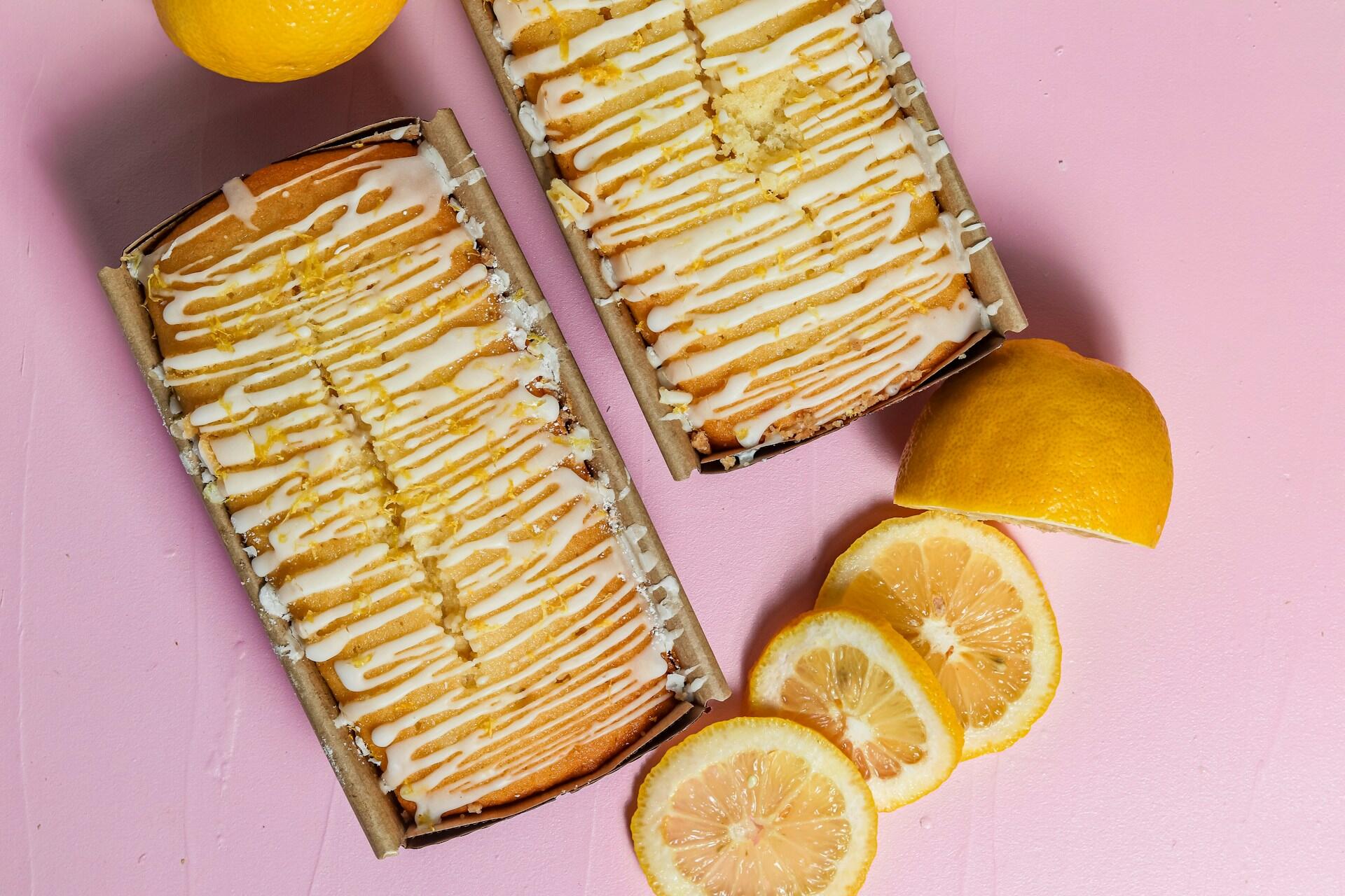 Two lemon drizzle cakes in brown paper trays sit on a pink surface. 