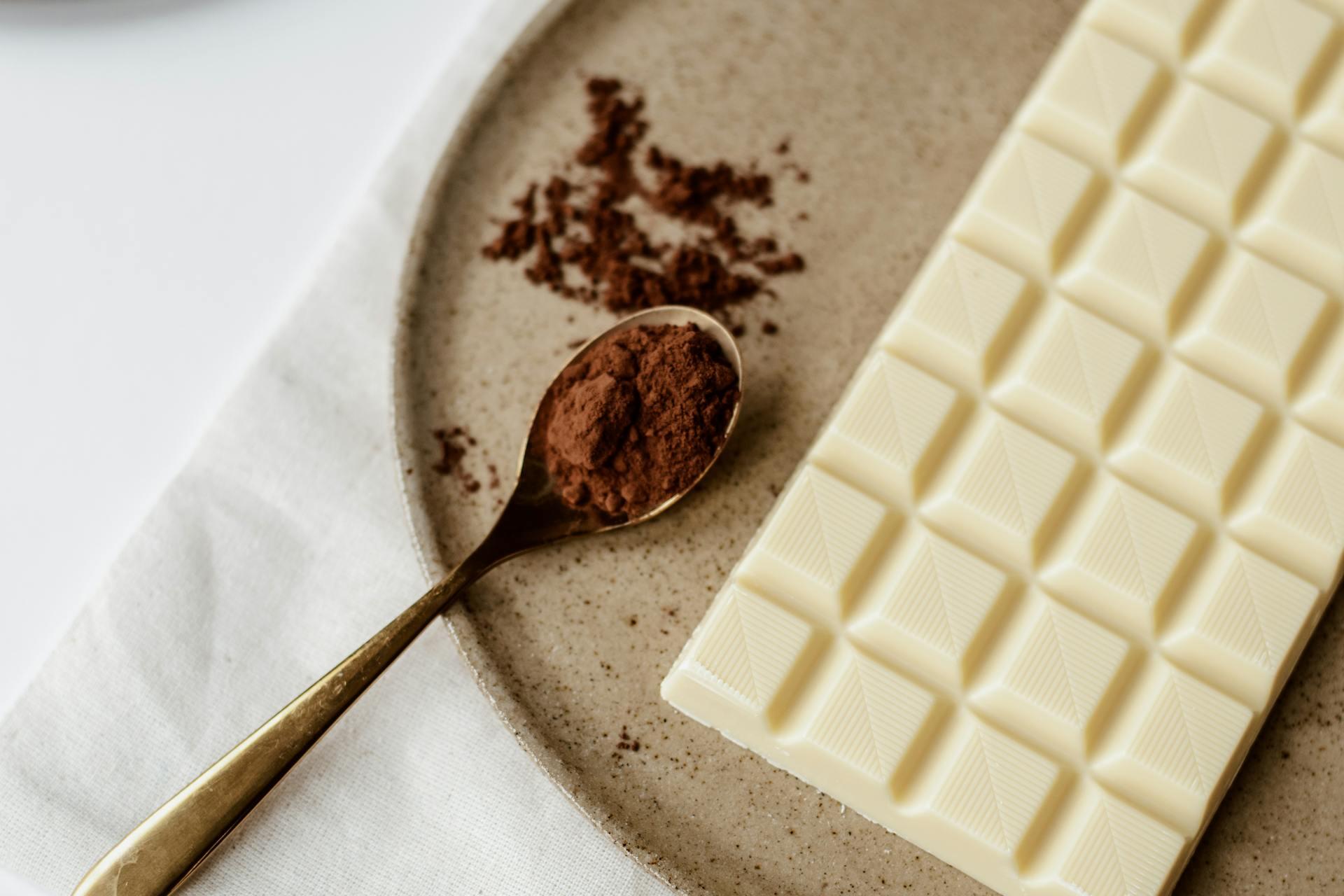 A white chocolate bar on a ceramic plate with a grid pattern, alongside a gold spoon holding cocoa powder.