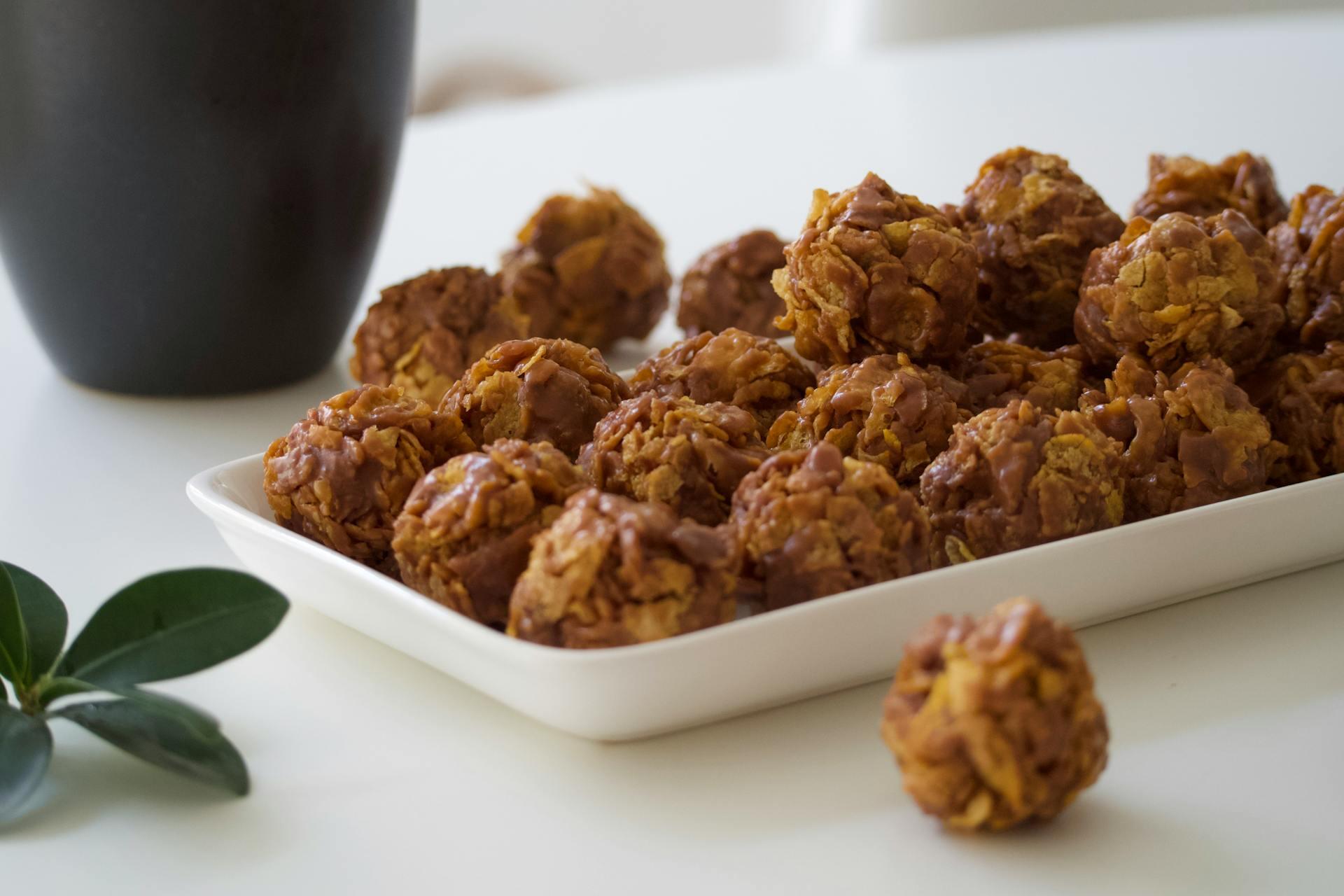 A white plate filled with clusters of cocoa cornflake cookies on a white table. A black mug and green leaves are nearby, creating a cozy atmosphere.