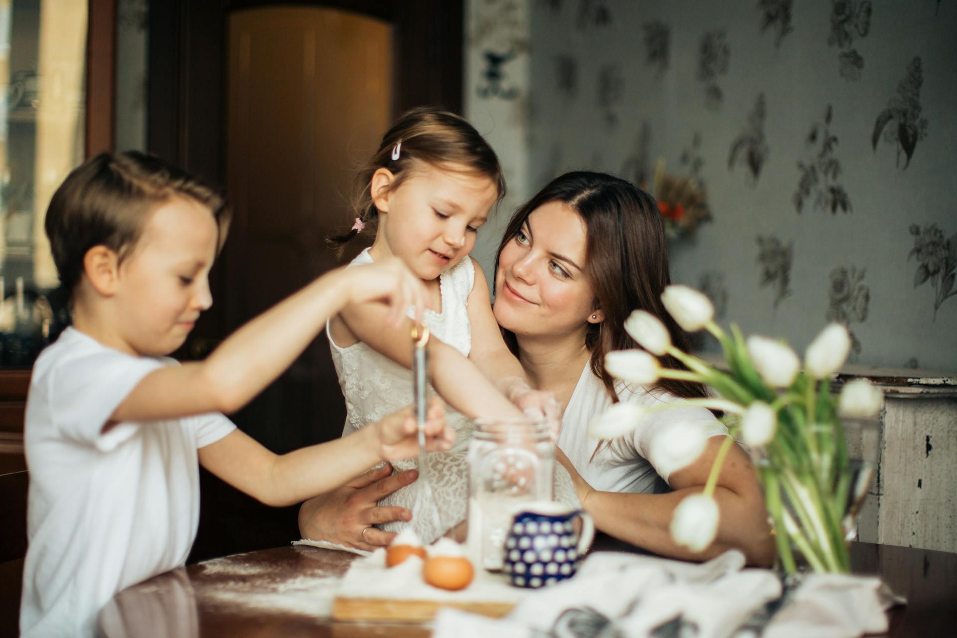 A mother and two children bake together at a kitchen table. The mother smiles warmly at them. Tulips, flour, and eggs enhance the cozy atmosphere.