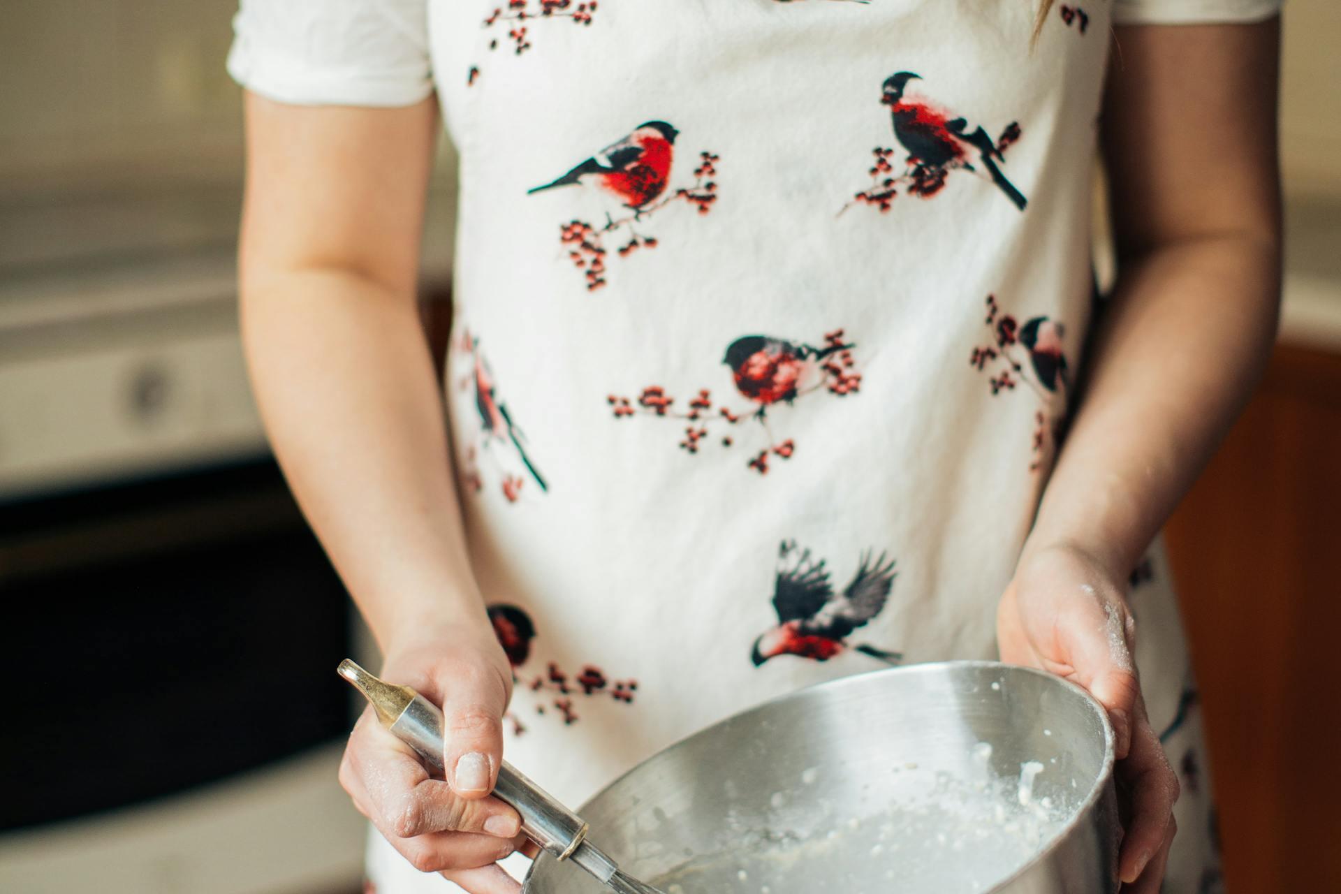 A person wearing a white apron with red and black bird designs holds a metal mixing bowl.