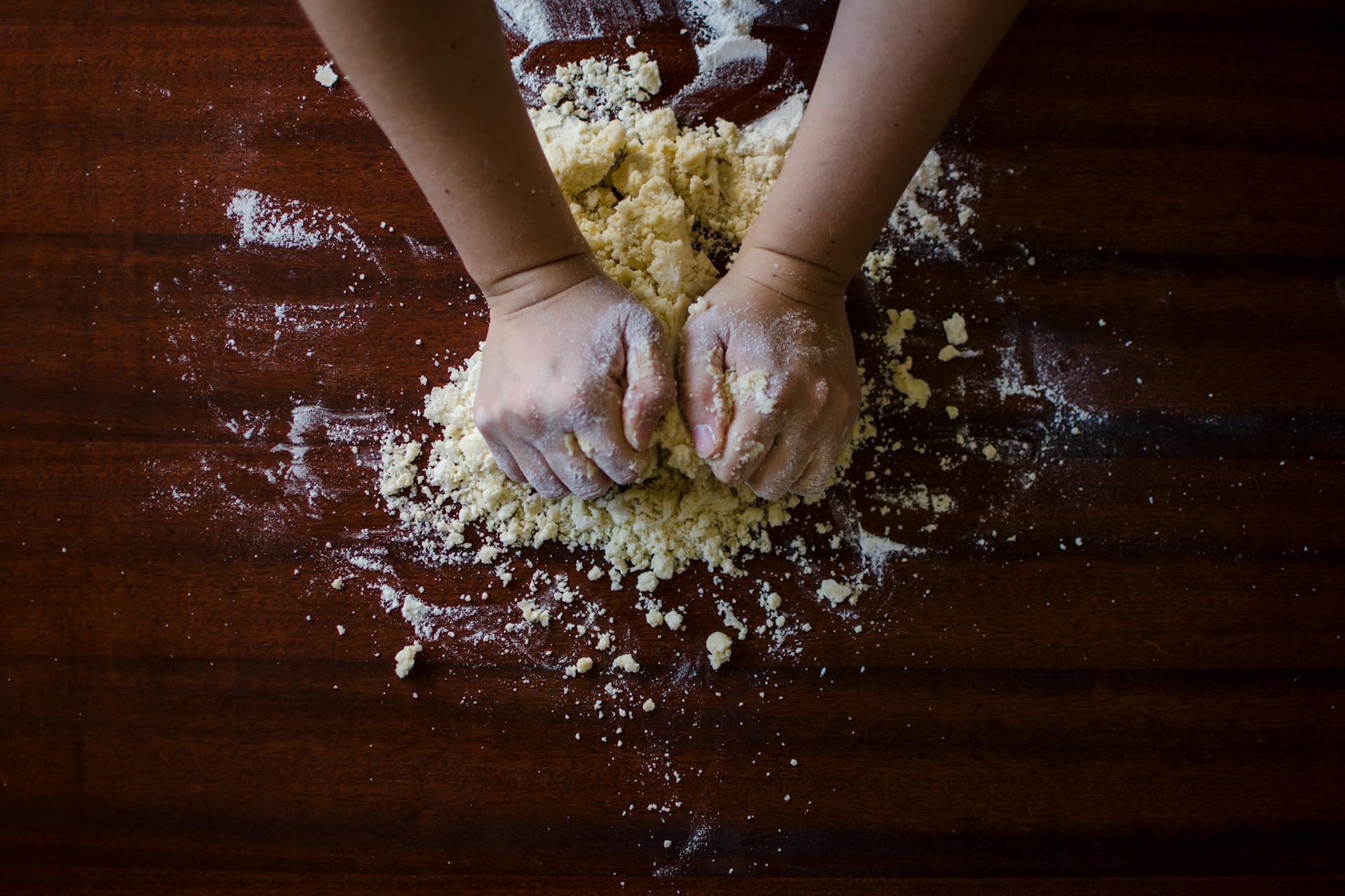 Hands kneading dough on a wooden table. Flour is scattered around, creating a feeling of homemade cooking and tactile engagement.