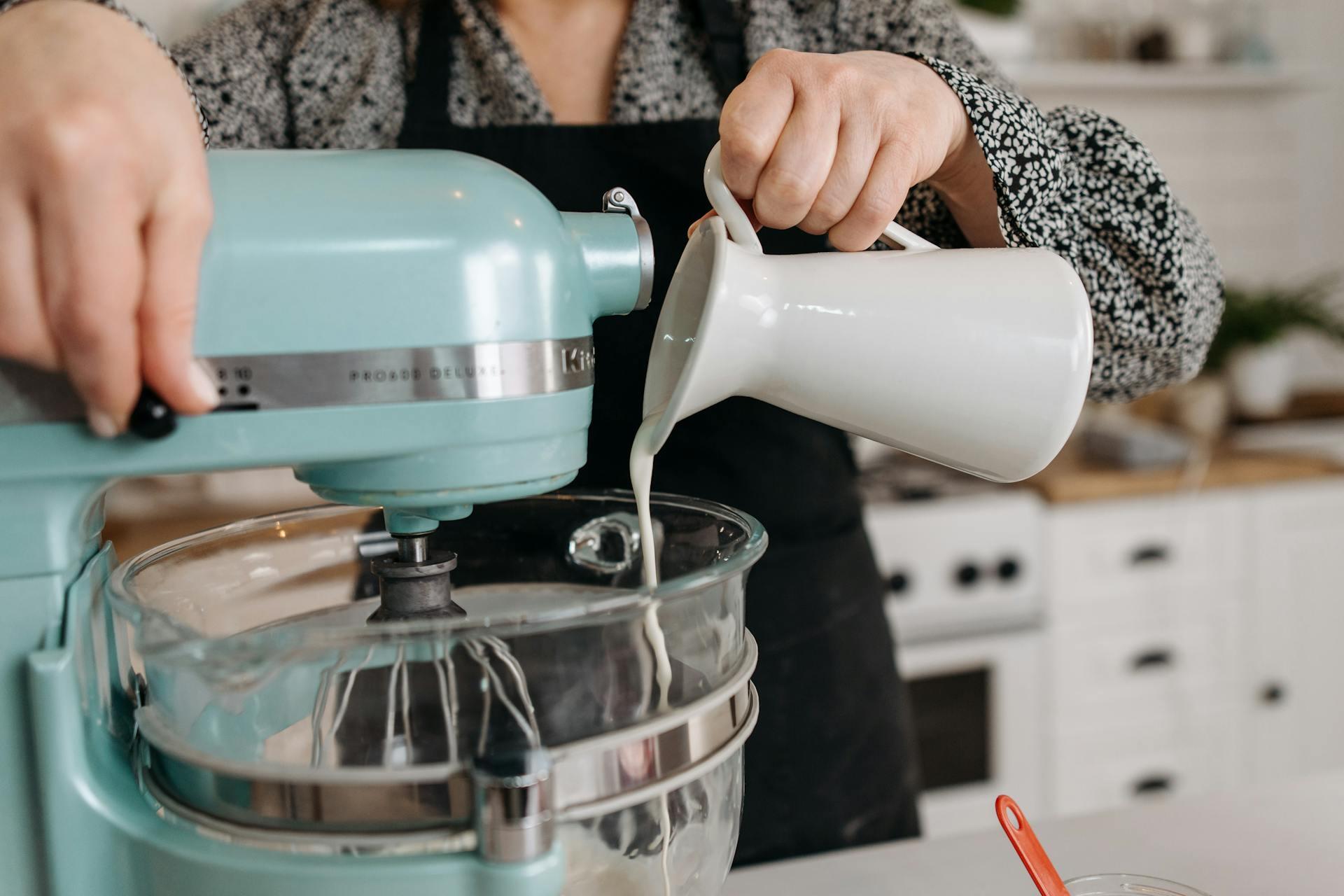 A person pours milk from a white pitcher into a blue stand mixer in a kitchen. 