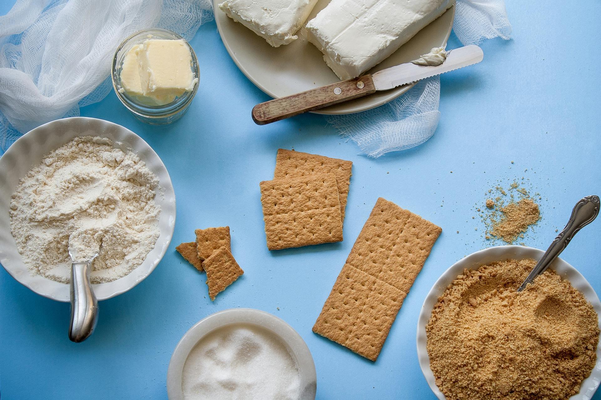 Ingredients on a blue surface include cream cheese, graham crackers, butter, flour, and sugar.