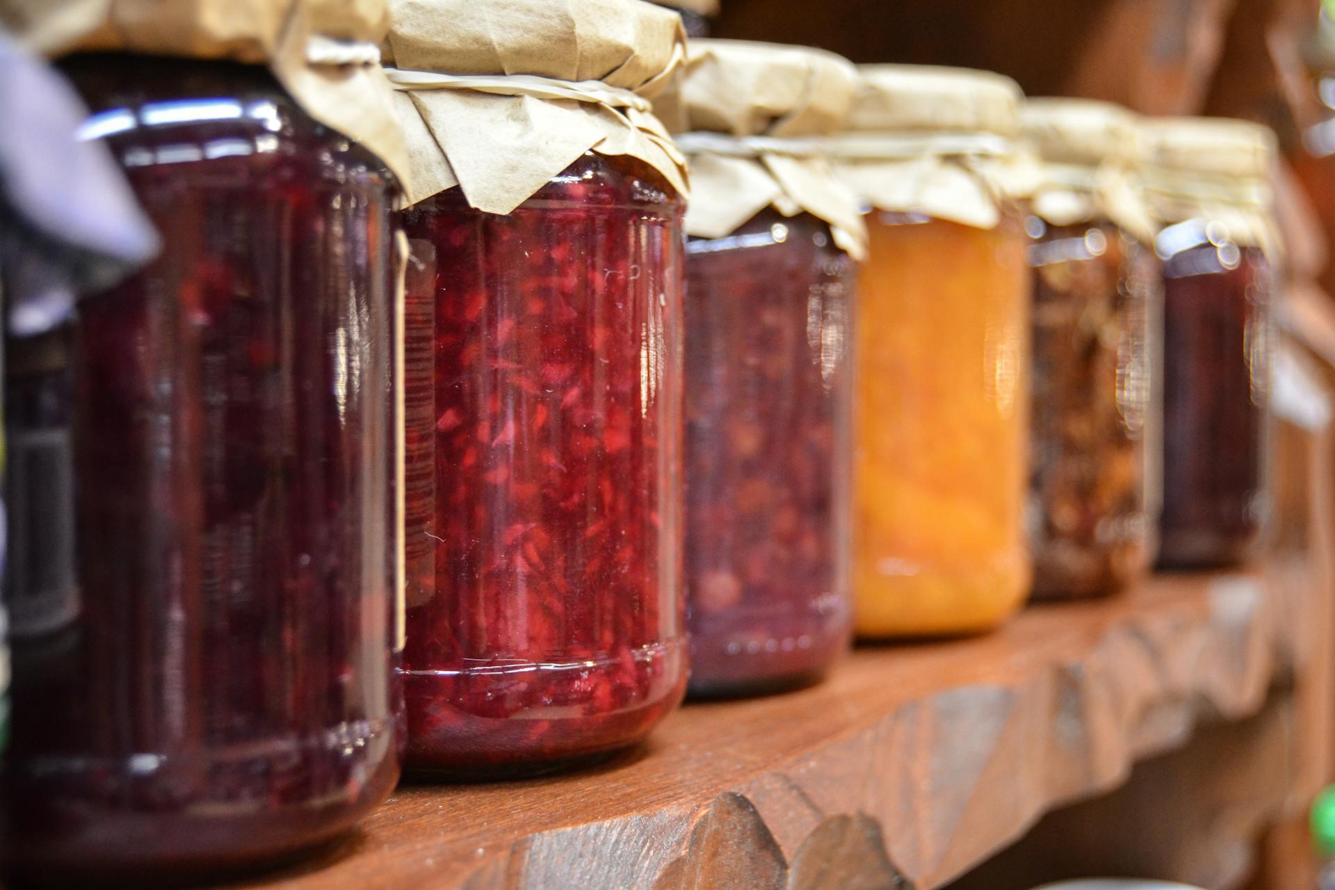 Close-up of a wooden shelf lined with various jars of preserves. Each jar has a paper cover and contains different brightly colored jams. Cozy, rustic setting.