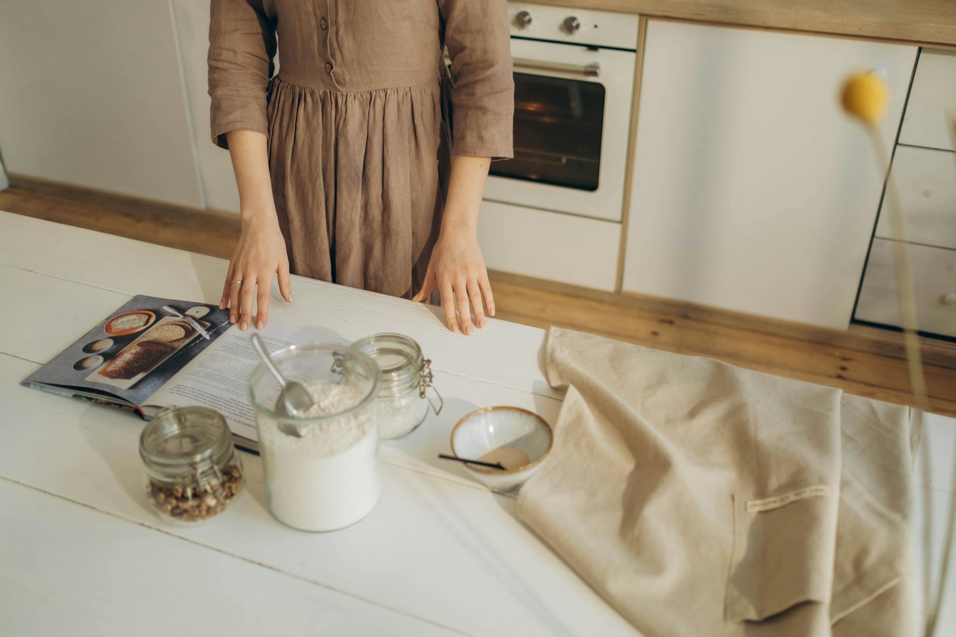 A person in a brown dress stands at a kitchen counter with an open cookbook, jars of flour and nuts, a bowl, and a beige apron, conveying a cozy baking scene.