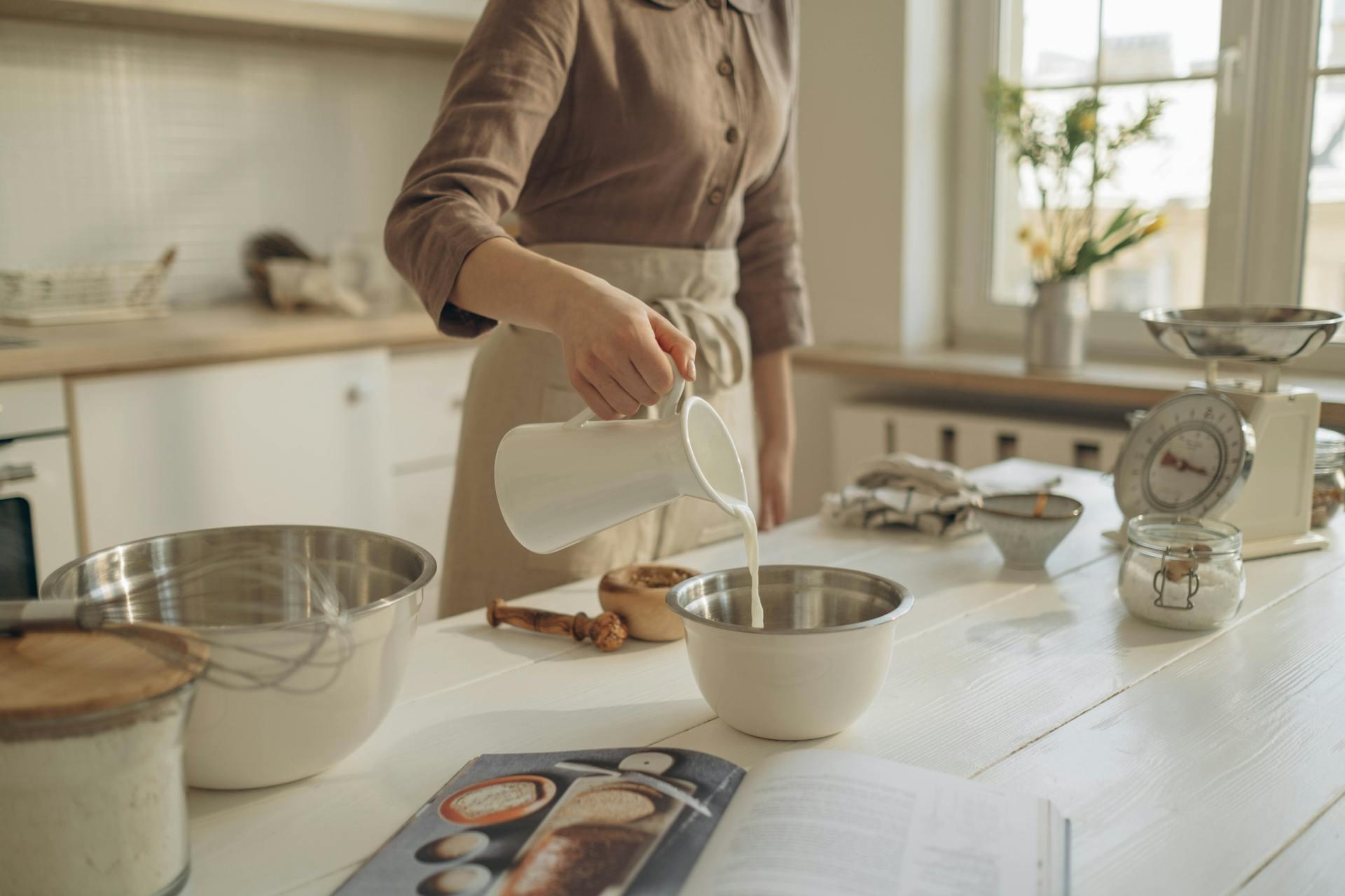 A person pours milk from a white jug into a metal bowl on a sunlit kitchen counter. Nearby, there are baking tools, a scale, and an open cookbook.