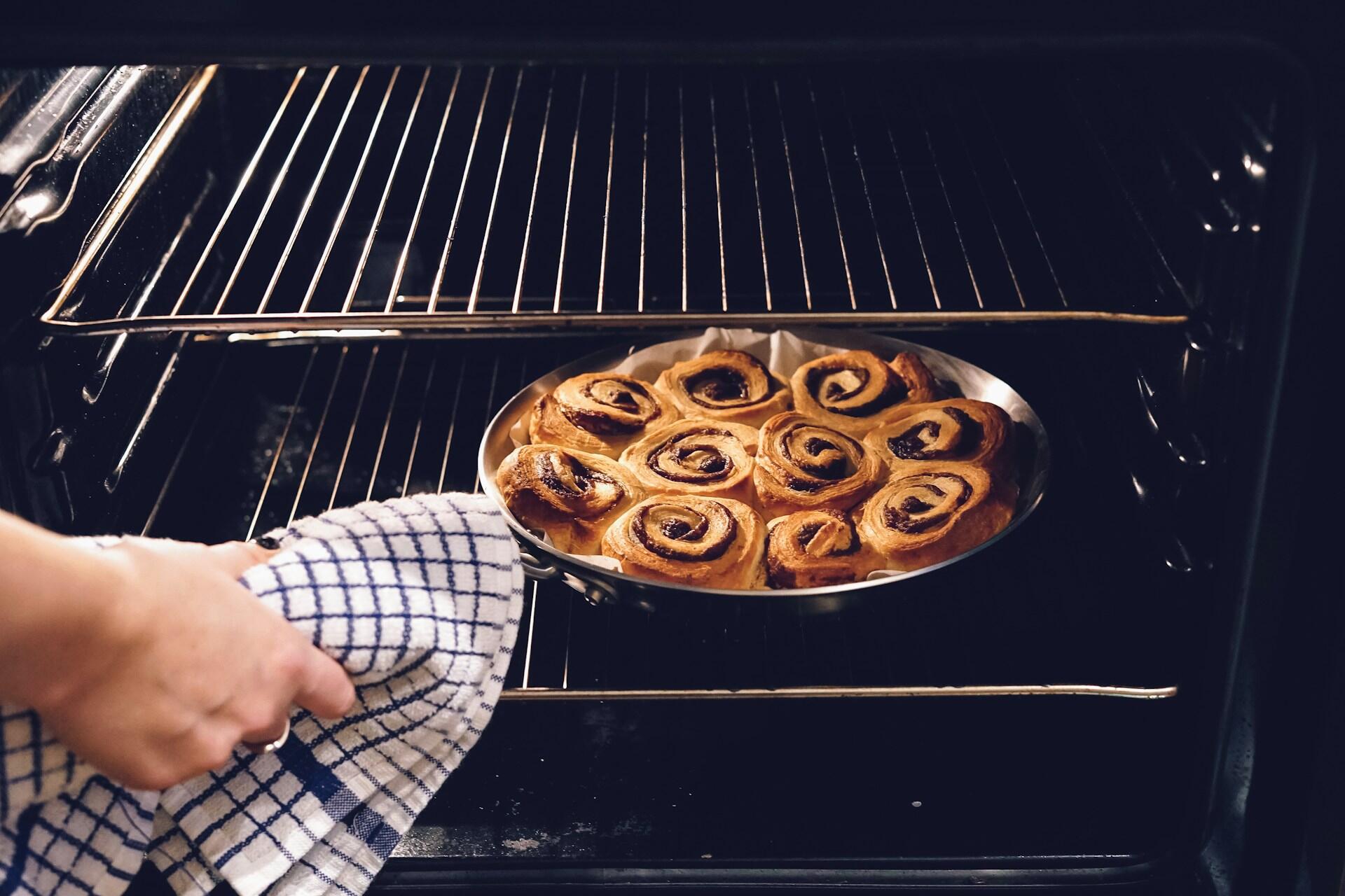 A person uses a checkered towel to remove a tray of freshly baked cinnamon rolls from an oven, conveying warmth and a sense of cozy home baking.